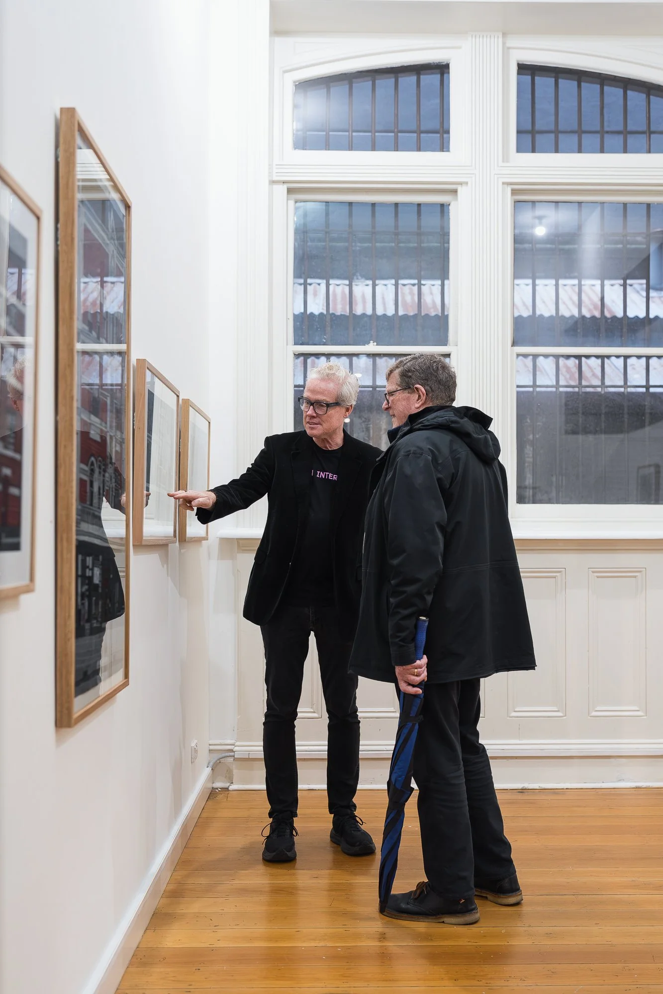 Two men in dark clothing viewing a series of framed photographic prints in a gallery, near tall arched windows; one pointing at a print, the other holding a furled umbrella.