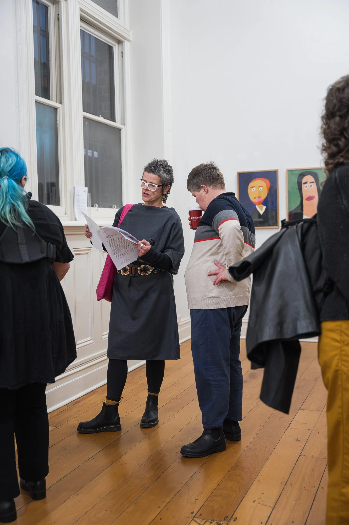 Woman reading from a handout to a young man holding a coffee cup, standing near two portrait paintings on the gallery wall.
