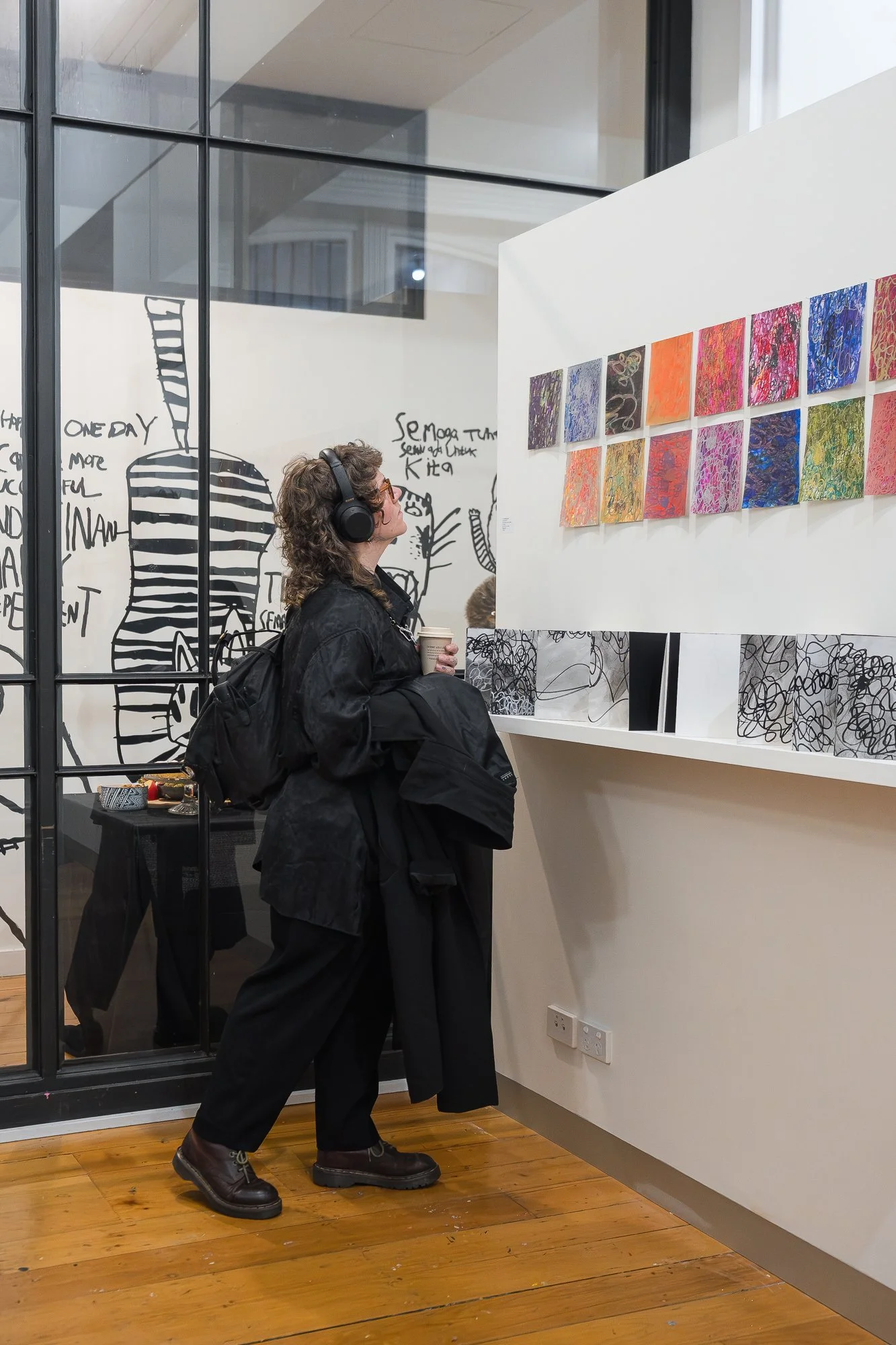 A visitor in all black with headphones looks up at a grid of vibrant small works, large black-and-white drawings visible behind her on the glass.