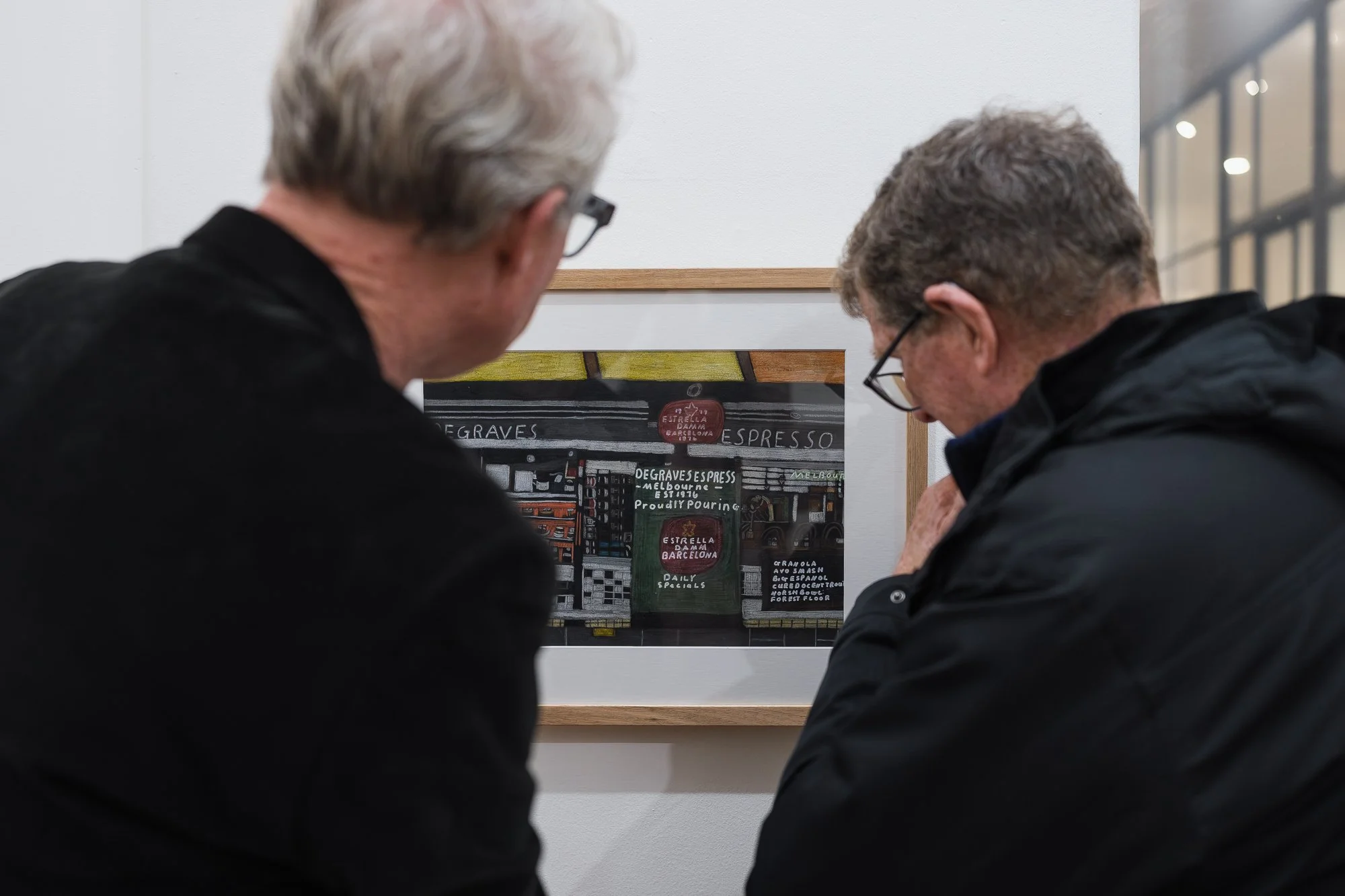 Close-up over the shoulders of two men examining a framed illustration of "Degraves Espresso" shopfront signage, near a gallery window.