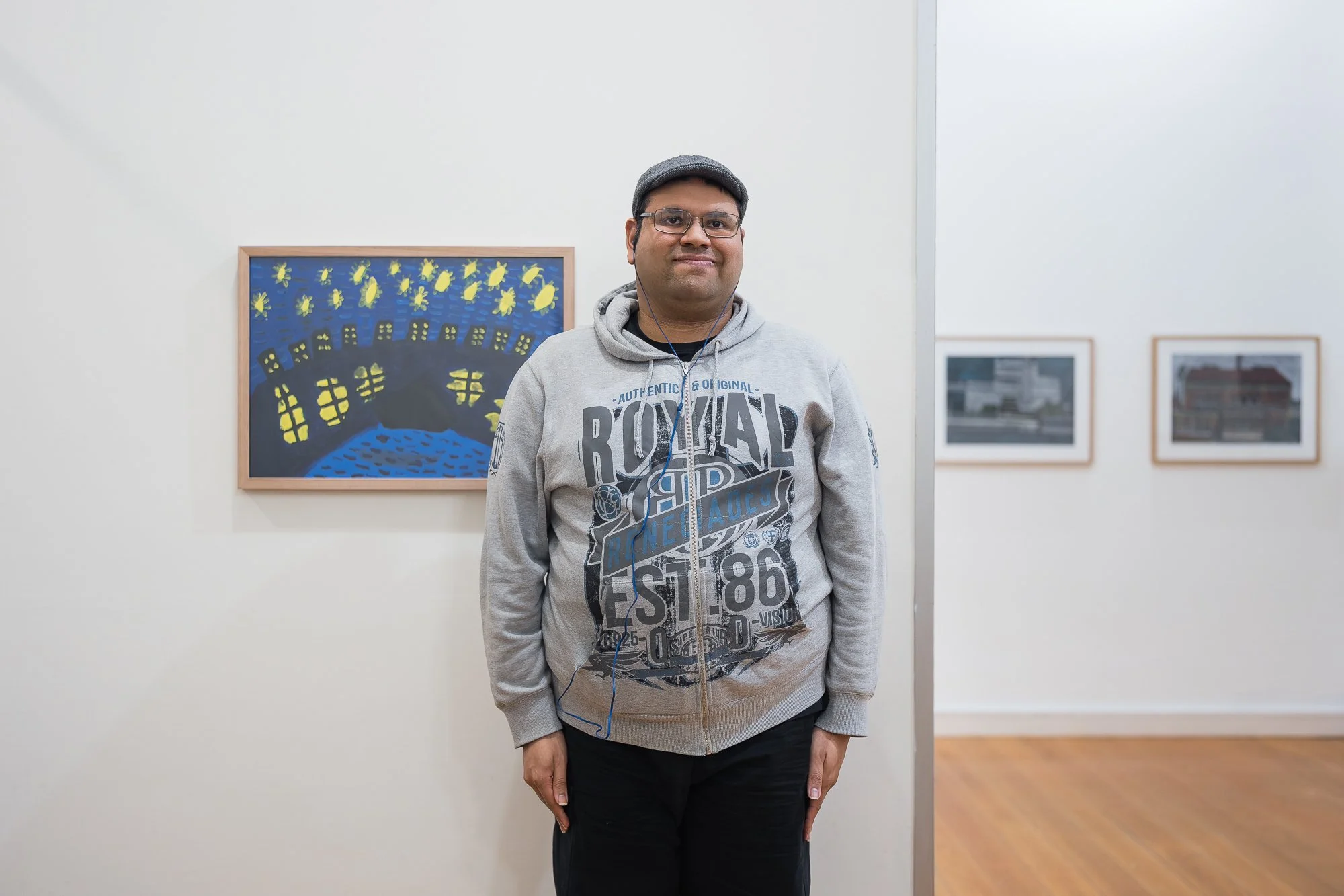 Portrait of a man in a grey "Royal Renegades" hoodie and flat cap, standing facing camera between two framed portrait paintings on the gallery wall.