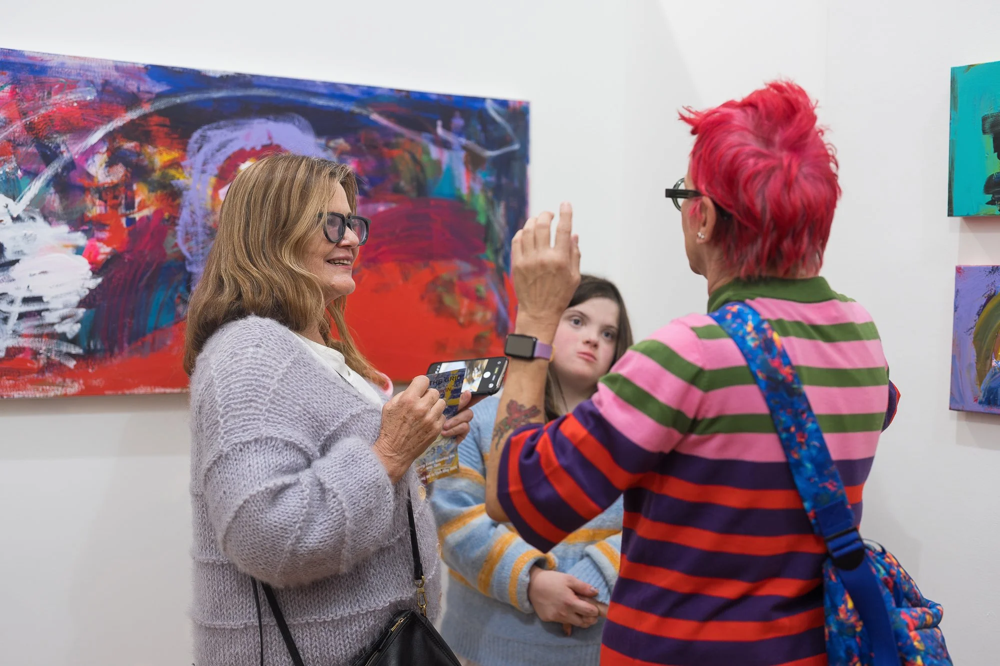 Three people chat animatedly in front of a large, vivid abstract painting in deep reds and blues.