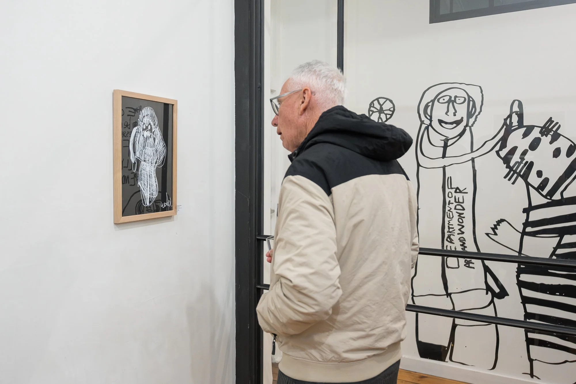 A visitor pauses to study a small framed work, surrounded by bold black-and-white figurative drawings on the gallery wall and window.