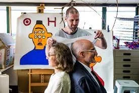 Artist measuring the height of a seated woman (left) and man, with a portrait of a person with six eyes in the background at Studio A, Sydney