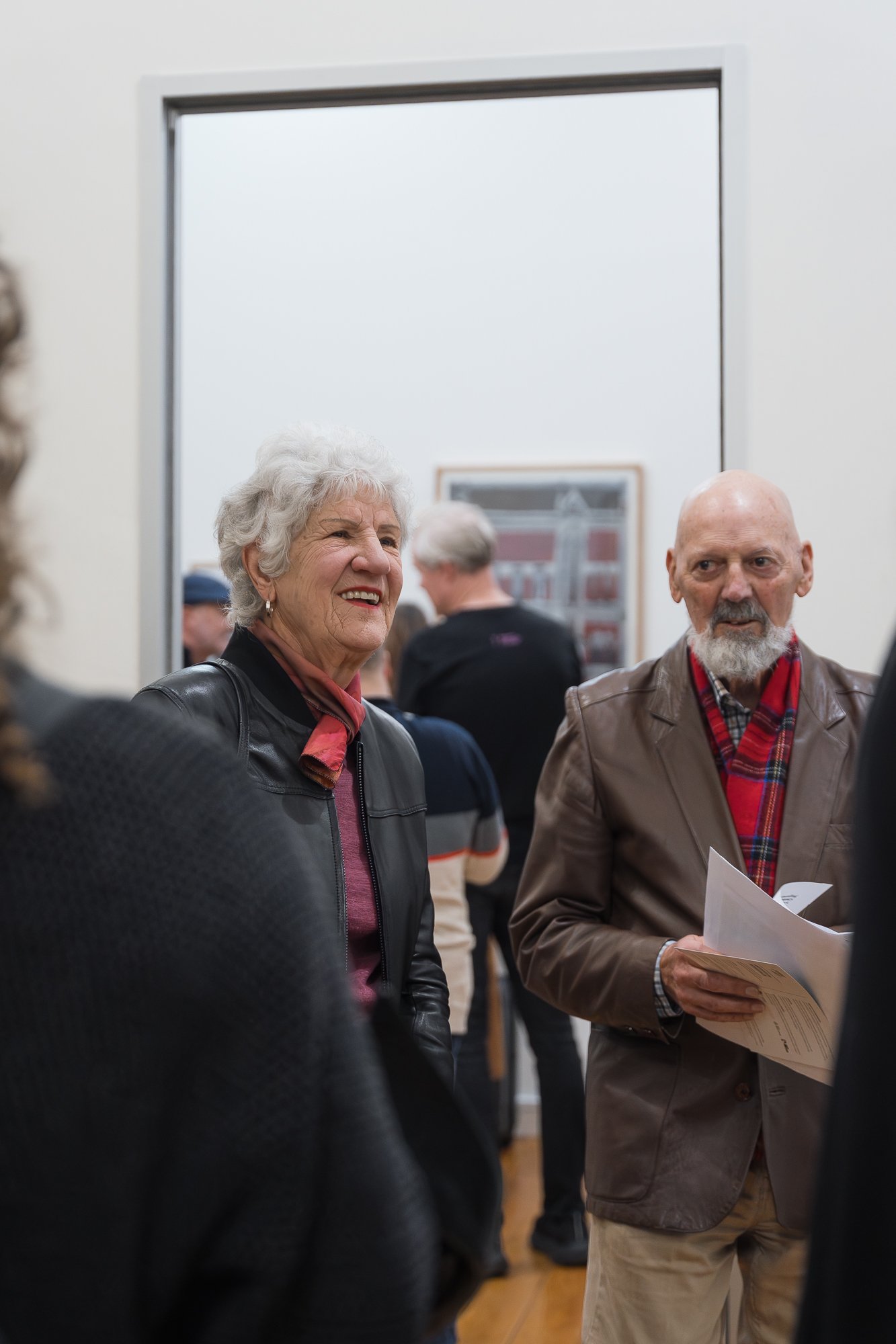 Close-up of an older woman and man in conversation, man holding an event program, framed building illustration blurred behind.