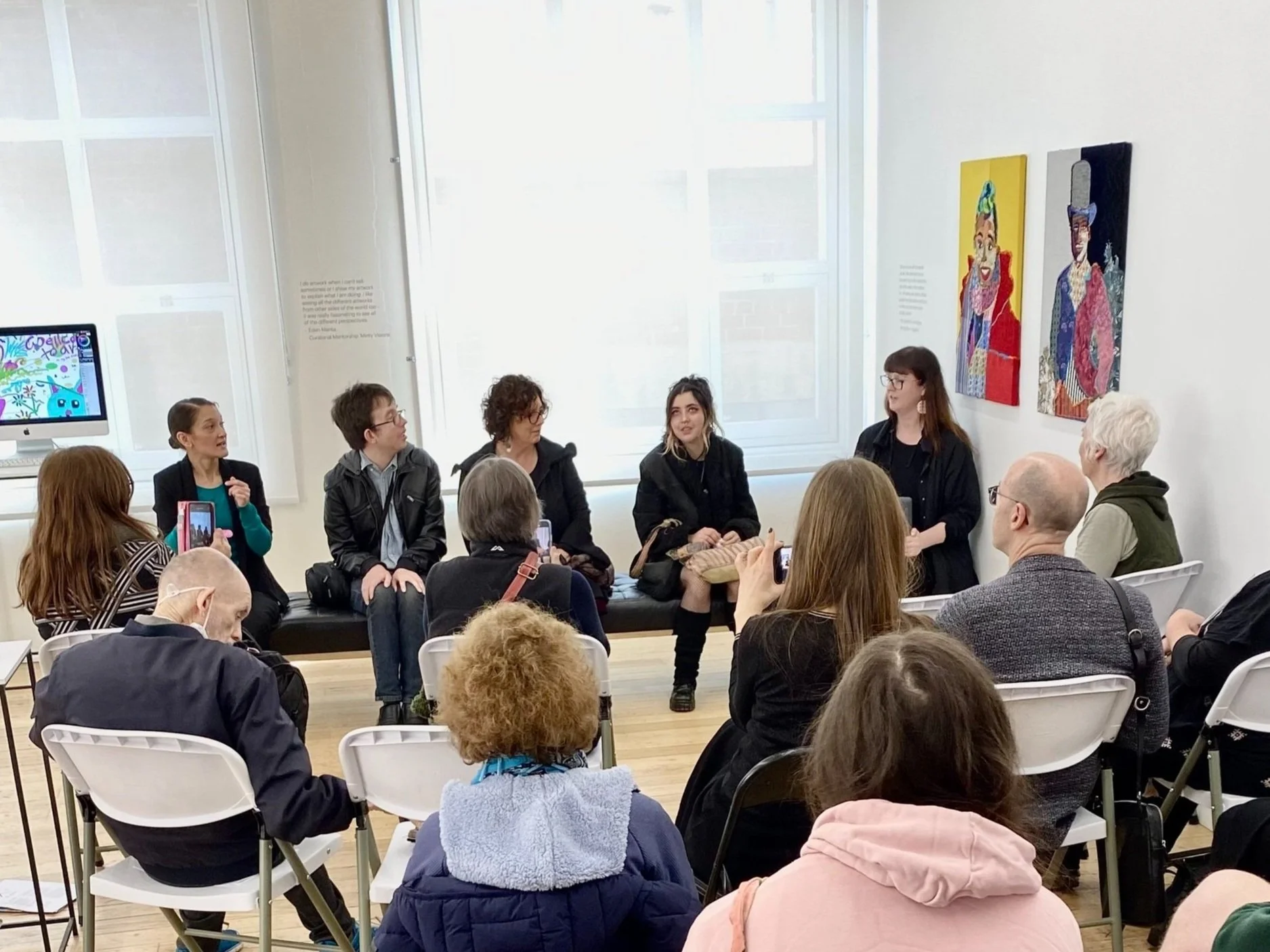 A group of people attending a panel discussion in an art gallery. The panelists are seated in front of an audience. The room has contemporary artwork displayed on the walls and a monitor showing digital art. The audience is sitting on white folding chairs, some taking pictures with mobile devices.
