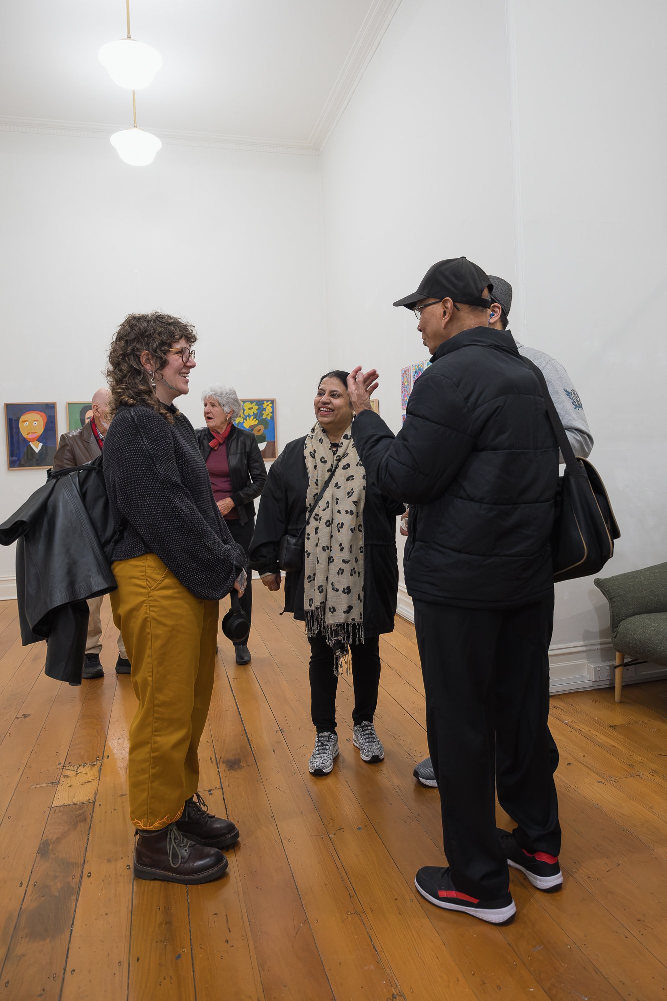 Four visitors in conversation in a gallery room, one woman in mustard trousers, portrait and floral paintings visible on the left wall.