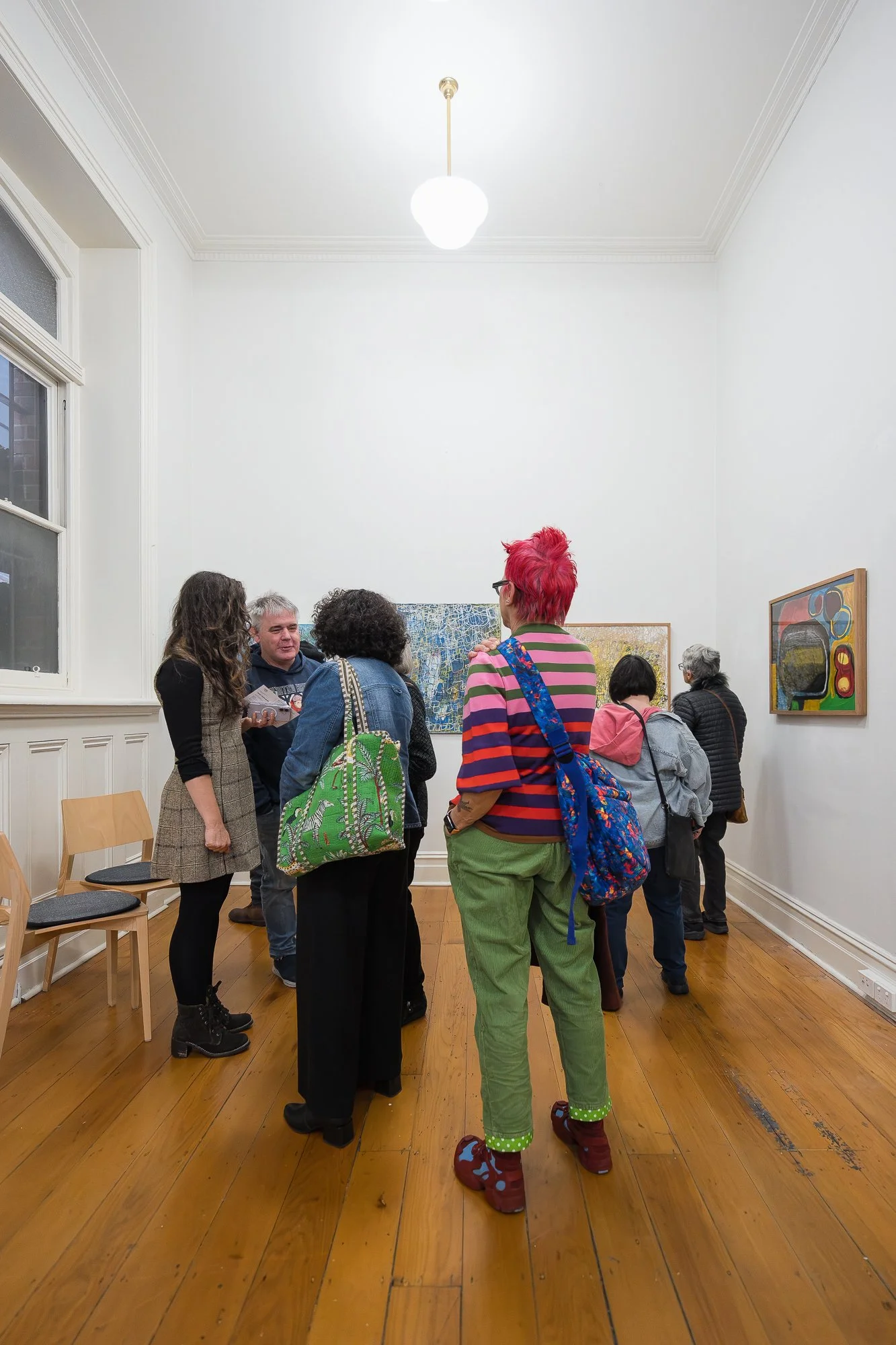 A group of visitors move through the gallery, chatting and taking in the works across two connected rooms.