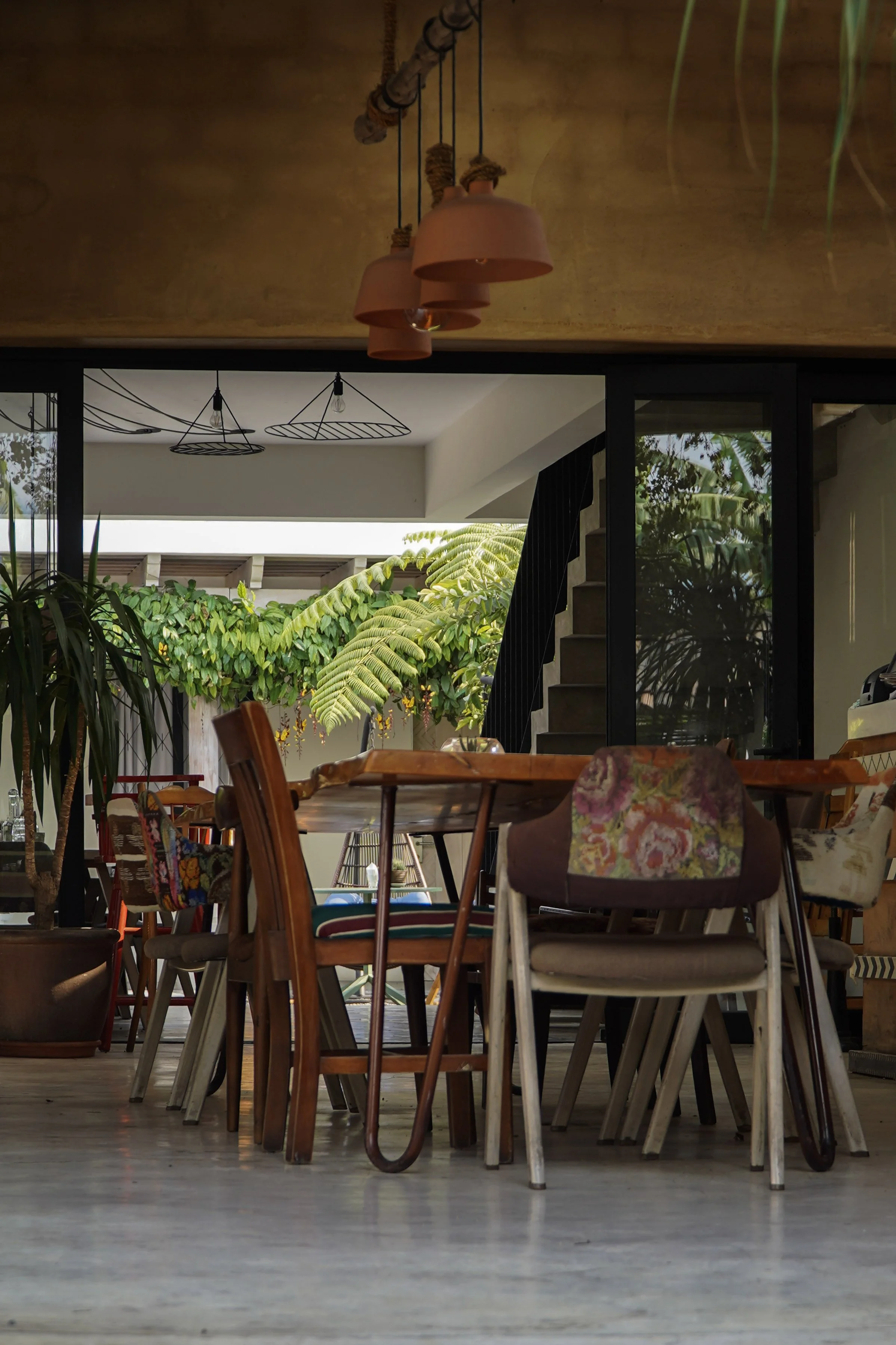 Interior view of a modern house with a dining area showing a variety of chairs around a wooden table, large sliding glass doors leading to an outdoor space, lush green plants, and contemporary pendant lighting.