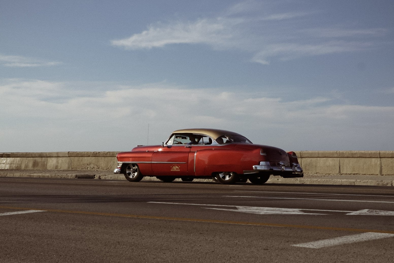 A vintage red and black classic car parked beside a concrete barrier on a road with a blue sky and light clouds in the background.