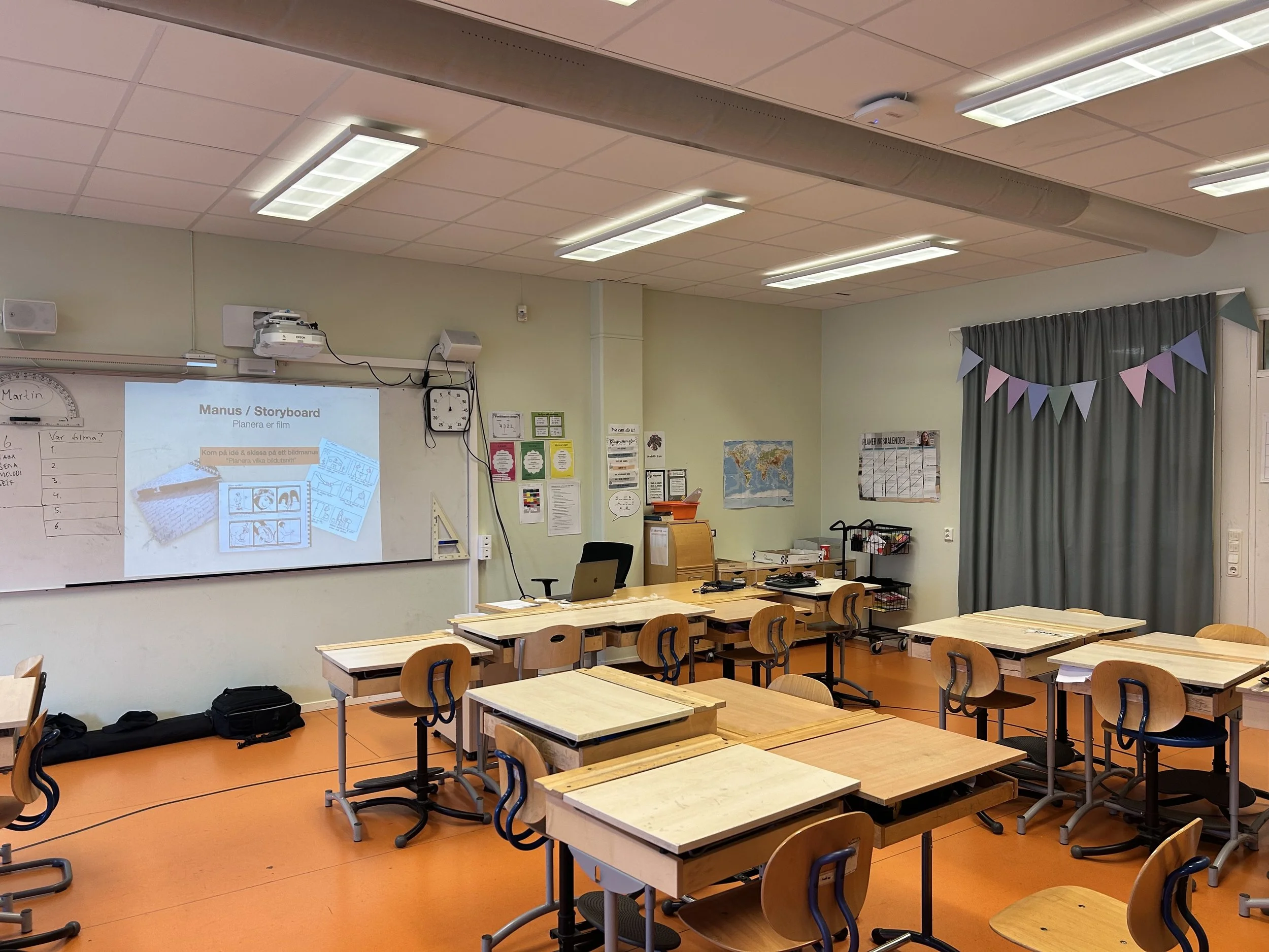 Ett tomt klassrum med runda träsittbord och stolar, en projektor och en whiteboard. Väggarna är ljusgröna, och det finns en blå, rosa och grön trekantig bår på en gardin.