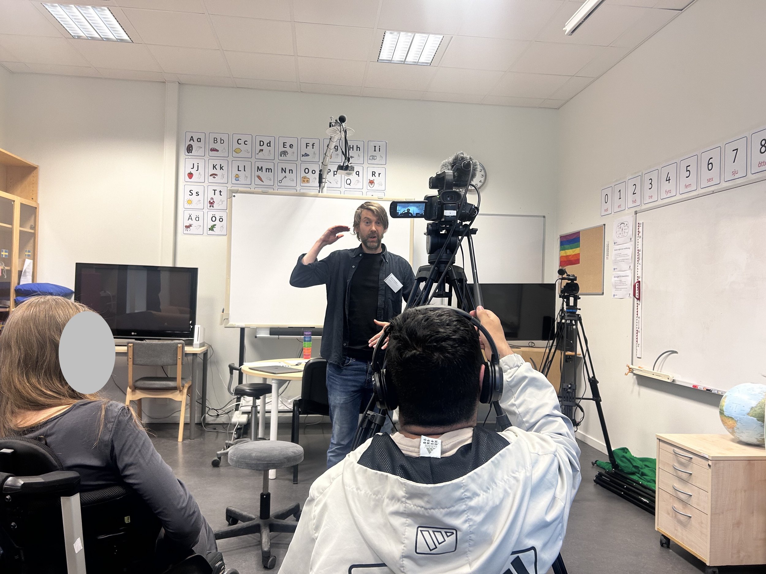 En man som talar eller förklarar något för en publik i ett klassrum, med en filmkamera riktad mot honom och elever som lyssnar. Klassrummet har skolväggar med alfabetet och siffror, samt en liten färgad flagga och en jordglob.