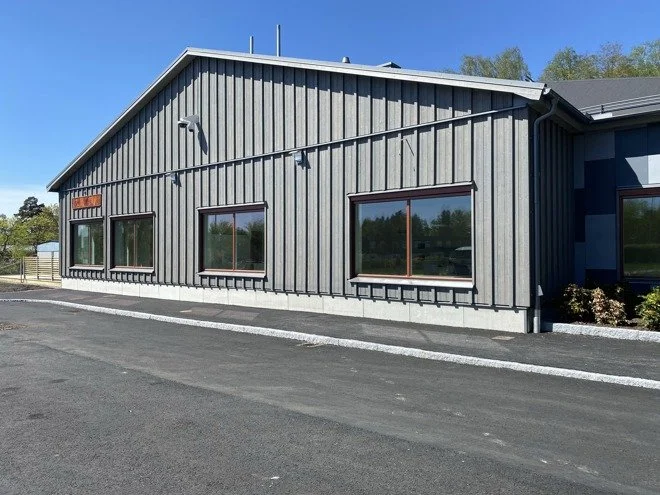 En modern byggnad med vertikala träpaneler och fyra stora fönster på framsidan, belägen vid en asfalterad väg och omgiven av grönskande träd.
