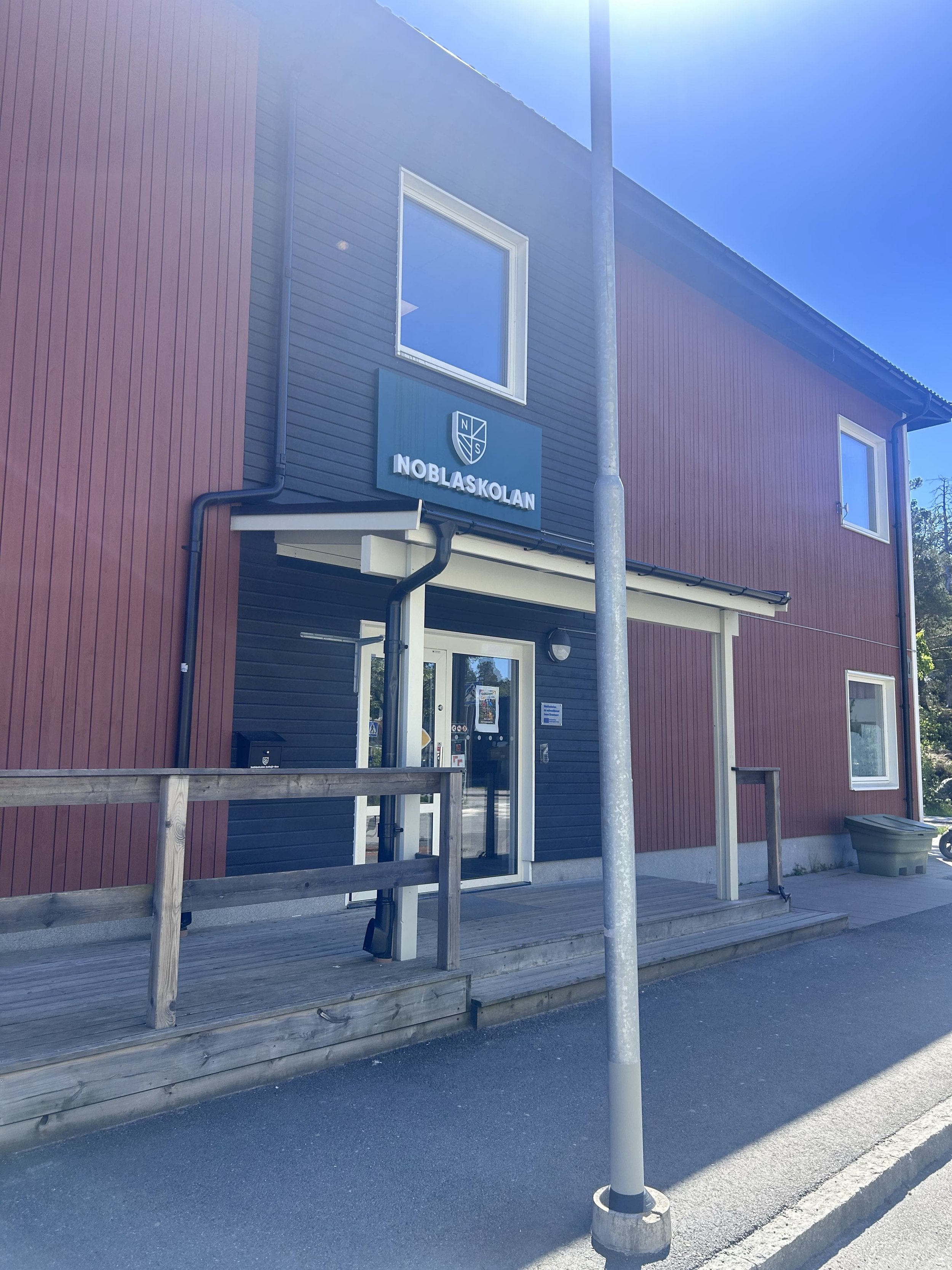 En modern tvåvåningsbyggnad med röda och mörkblå fasad, har en ingång med dörr och ett skylt som säger Noblaskolan.