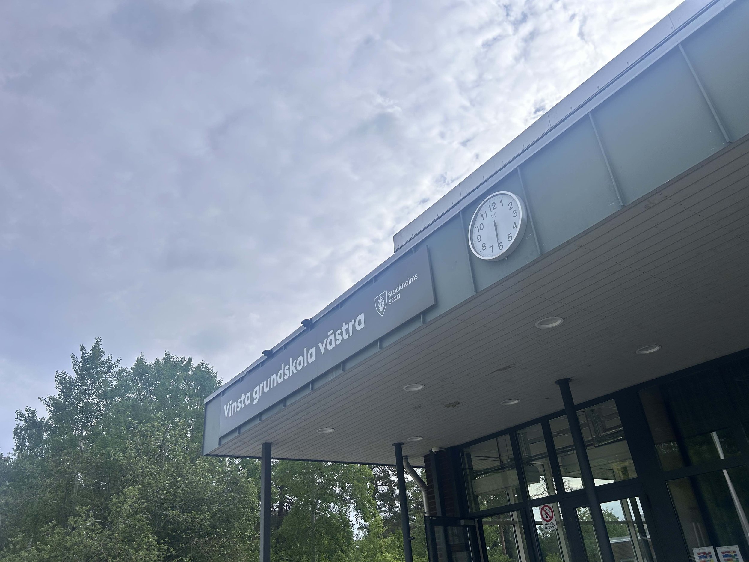 En modern skolbyggnad med en rund klocka på fasaden visar ungefär klockan 11:55, omgiven av gröna träd och en blå himmel.