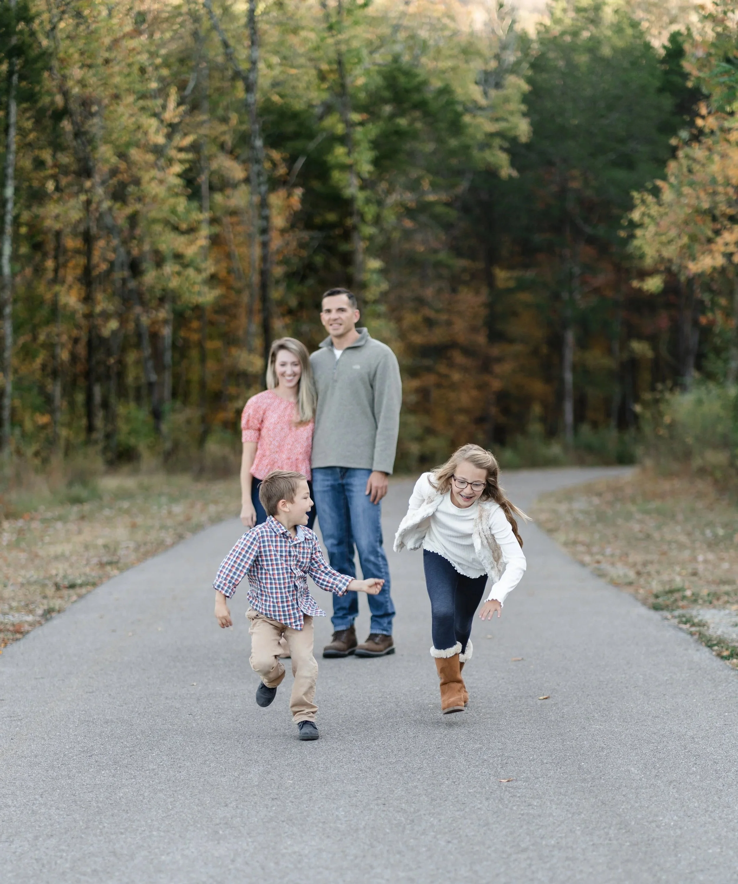 A family of five enjoying a fall day outdoors on a paved path surrounded by trees with colorful autumn leaves. The two children are playing and running, while the parents smile and watch.