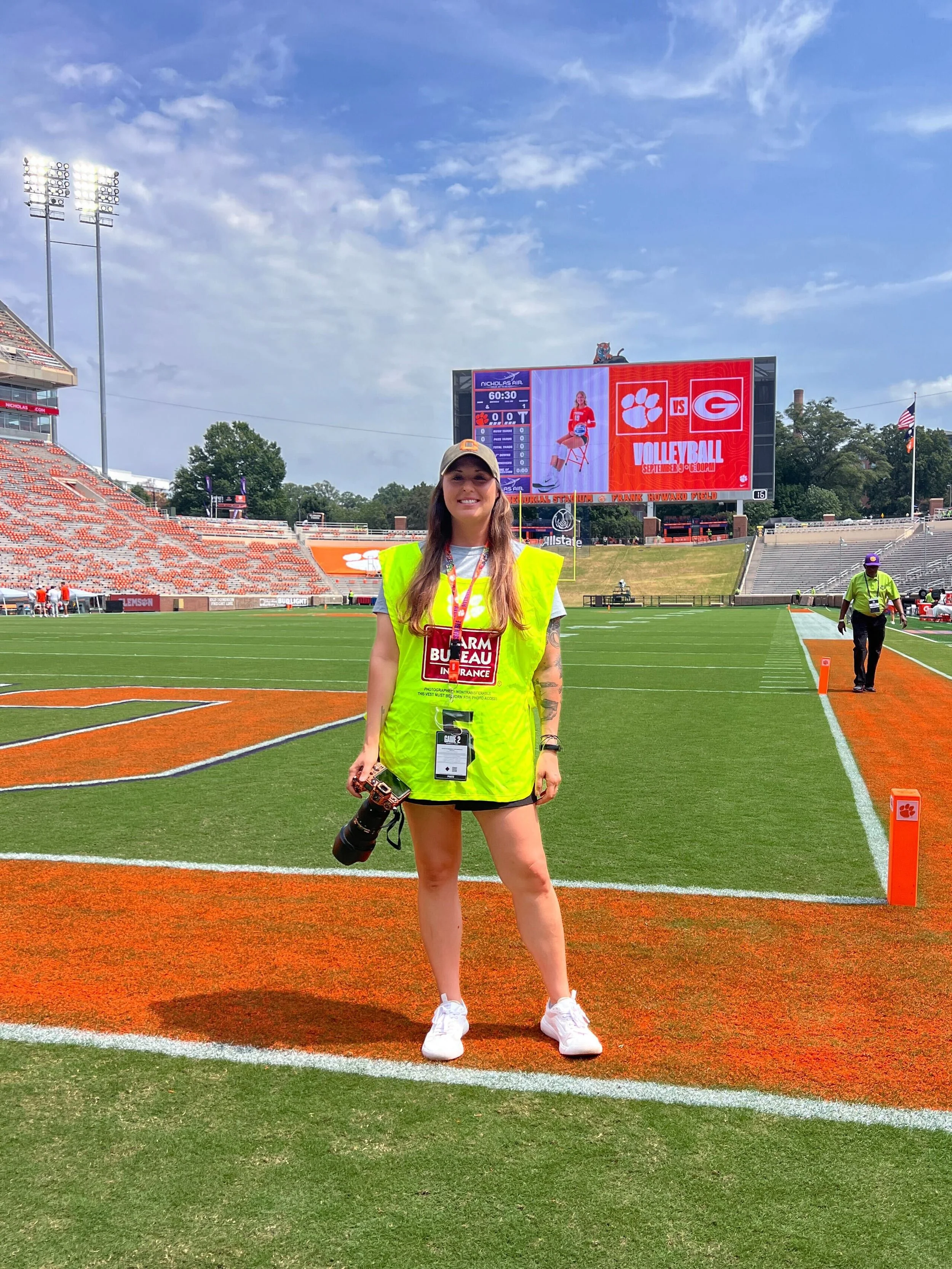 A woman standing on a football field with a camera in her hand, wearing a yellow safety vest and a gray cap, smiling, with a large digital scoreboard and empty stadium seats in the background.