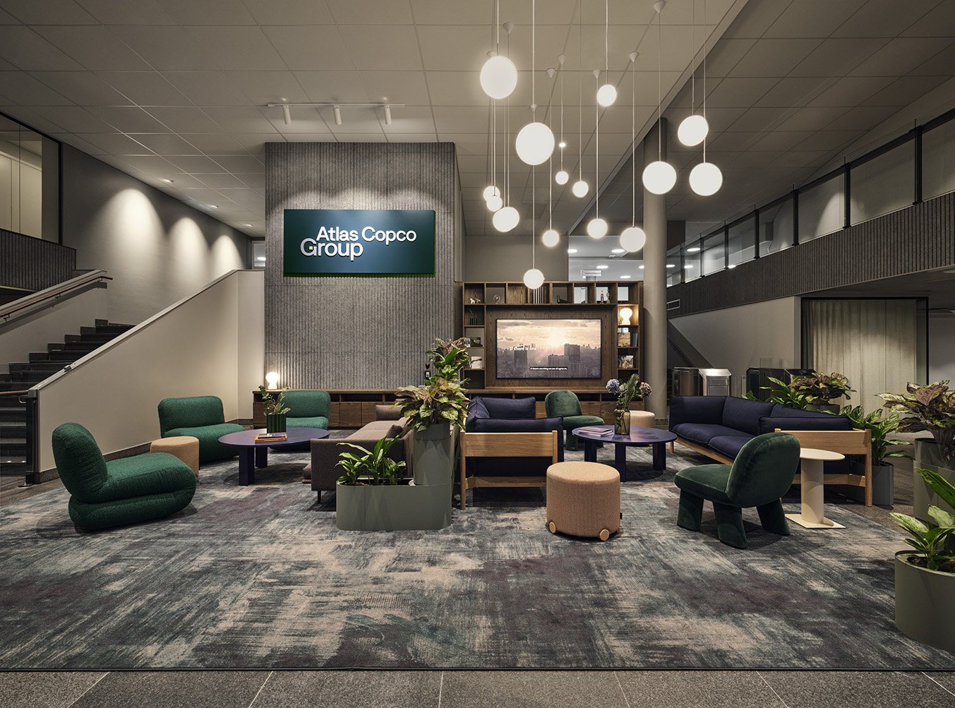 En modern lobby med mysiga soffor och fåtöljer, växter, ett stort tv-skärm, trappa, och ett signaltavla från Atlas Copco Group.