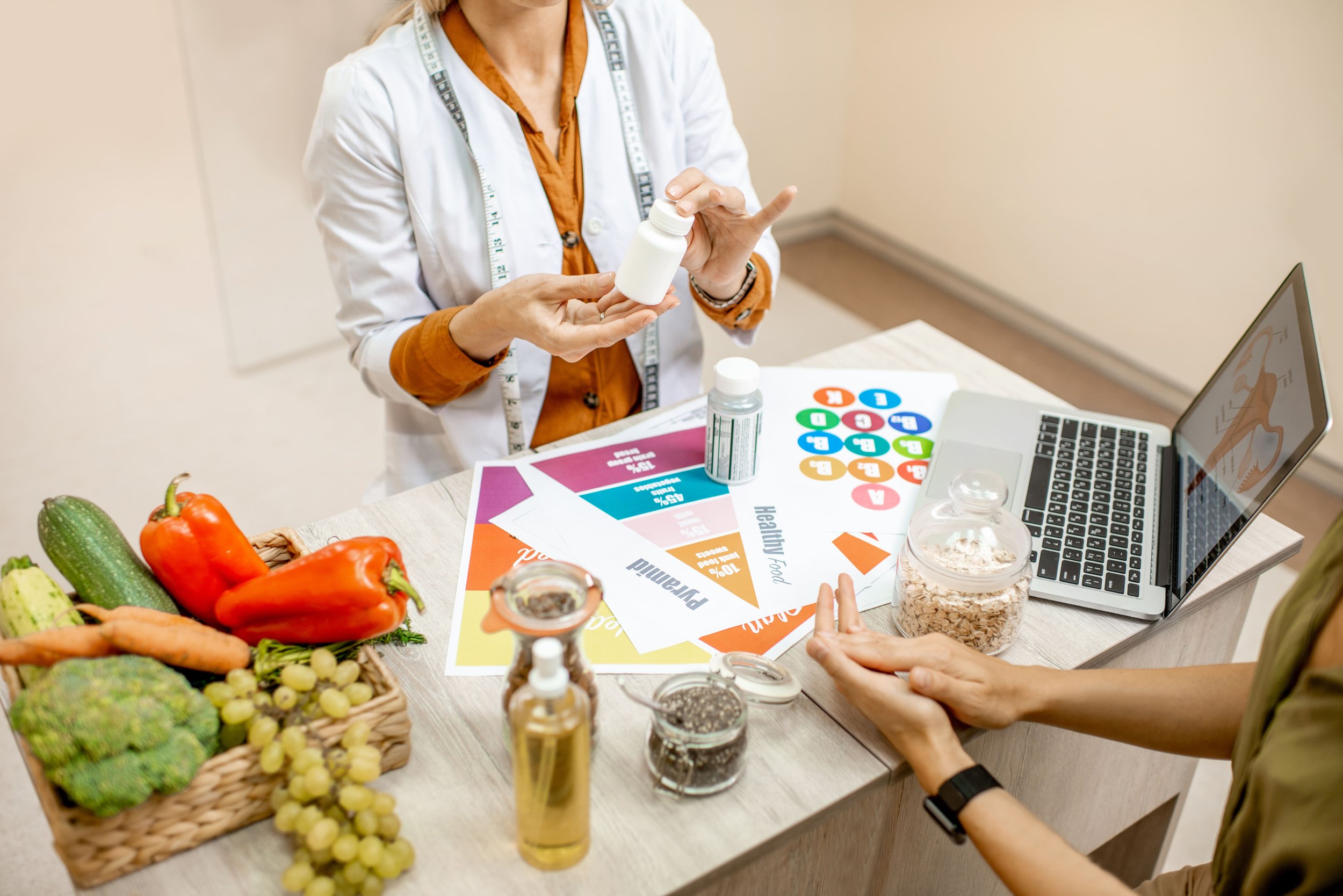 A person in a white coat and measuring tape is showing a supplement bottle, with another person seated at the desk, possibly in a consultation. The desk has fresh vegetables, informational pamphlets, a laptop, and jars of seeds or grains, indicating a health or nutrition planning session.
