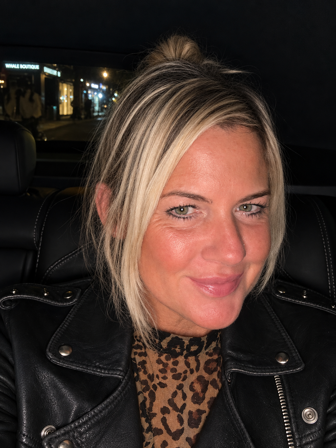 A woman with blonde hair and light green eyes sitting in a vehicle at night, wearing a black leather jacket and animal print top, with a city street visible in the background.