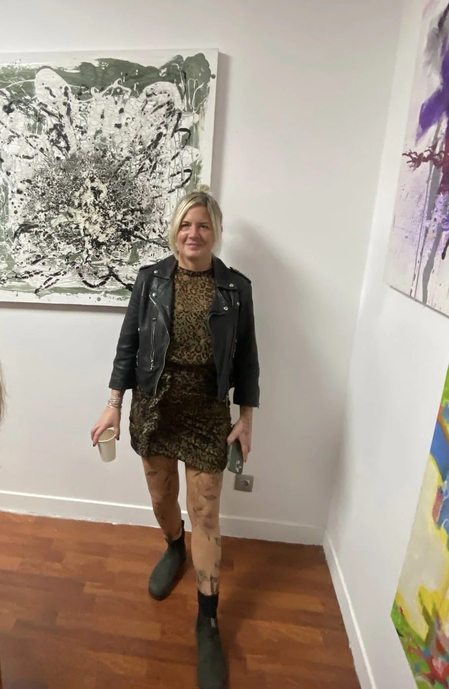 A woman with blonde hair wearing a black leather jacket, a leopard-print dress, and black boots standing in an art gallery with abstract paintings on the walls.