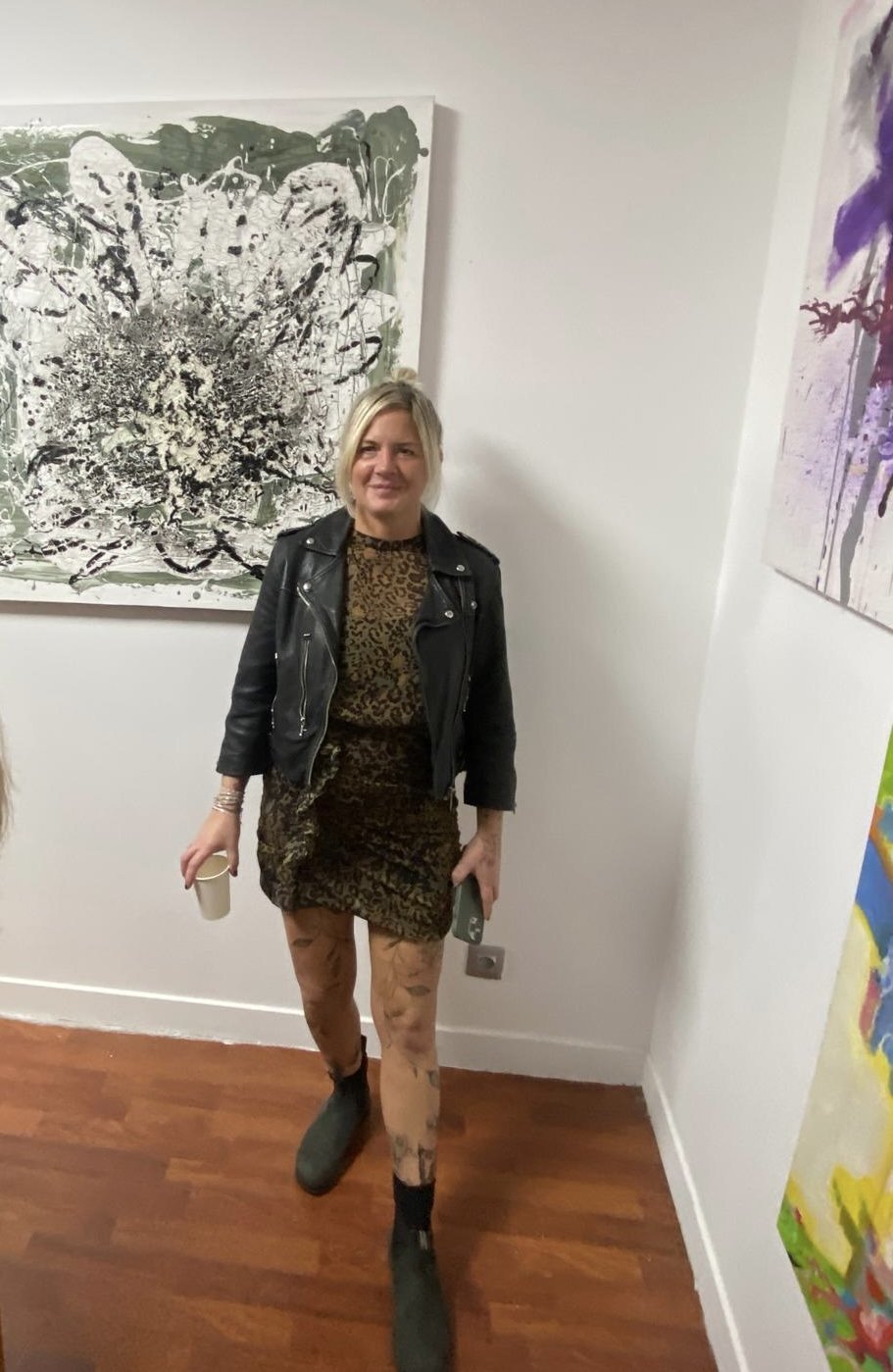 Woman with blonde hair wearing a black leather jacket, leopard print dress, tan patterned tights, and black shoes standing in an art gallery with abstract paintings on the wall.