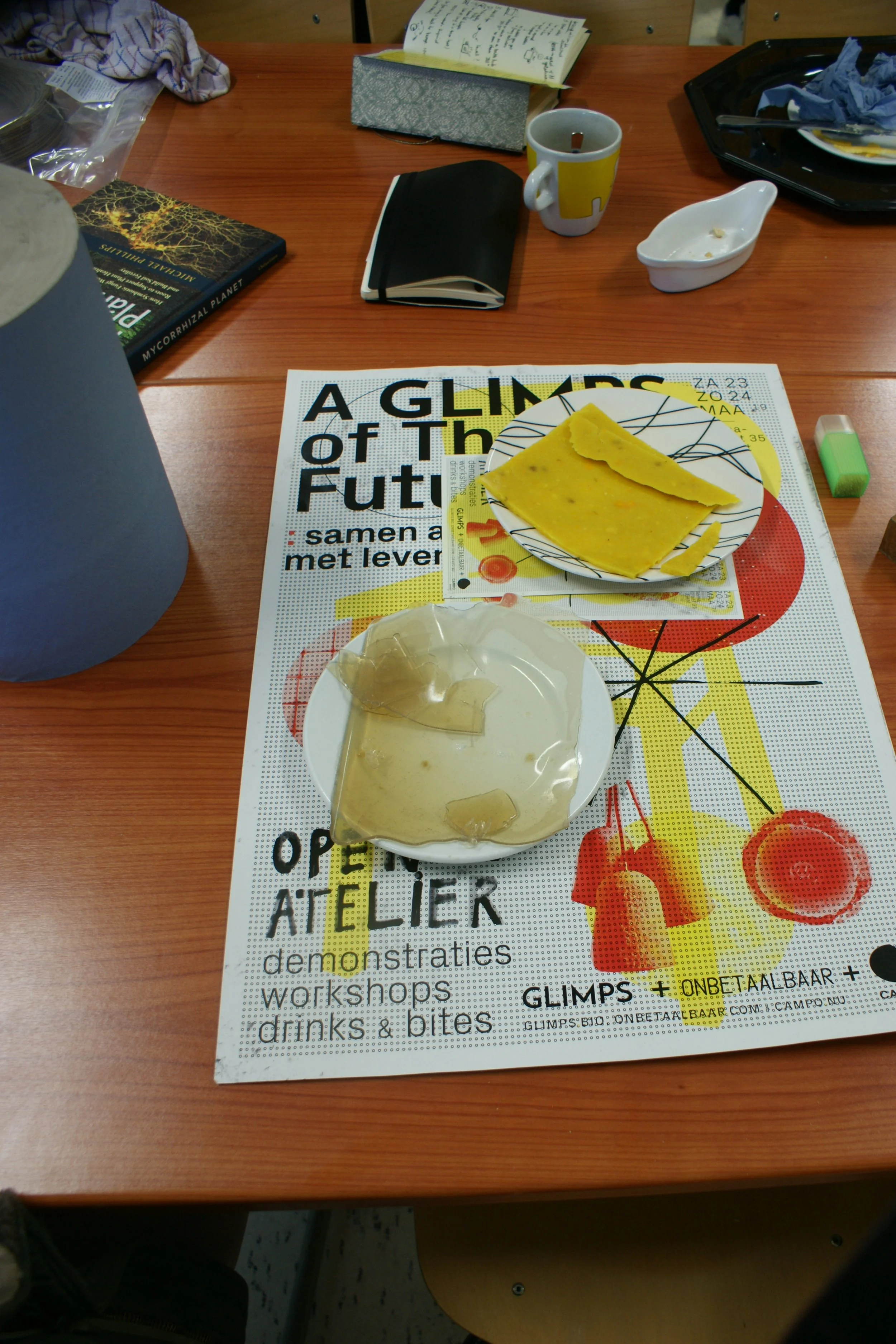 A table with a frequent magazine, a black notebook, a mug, a tissue box, some scraps of food, a glazed plate, and a plate with yellow crepes on a spread-out workshop poster.