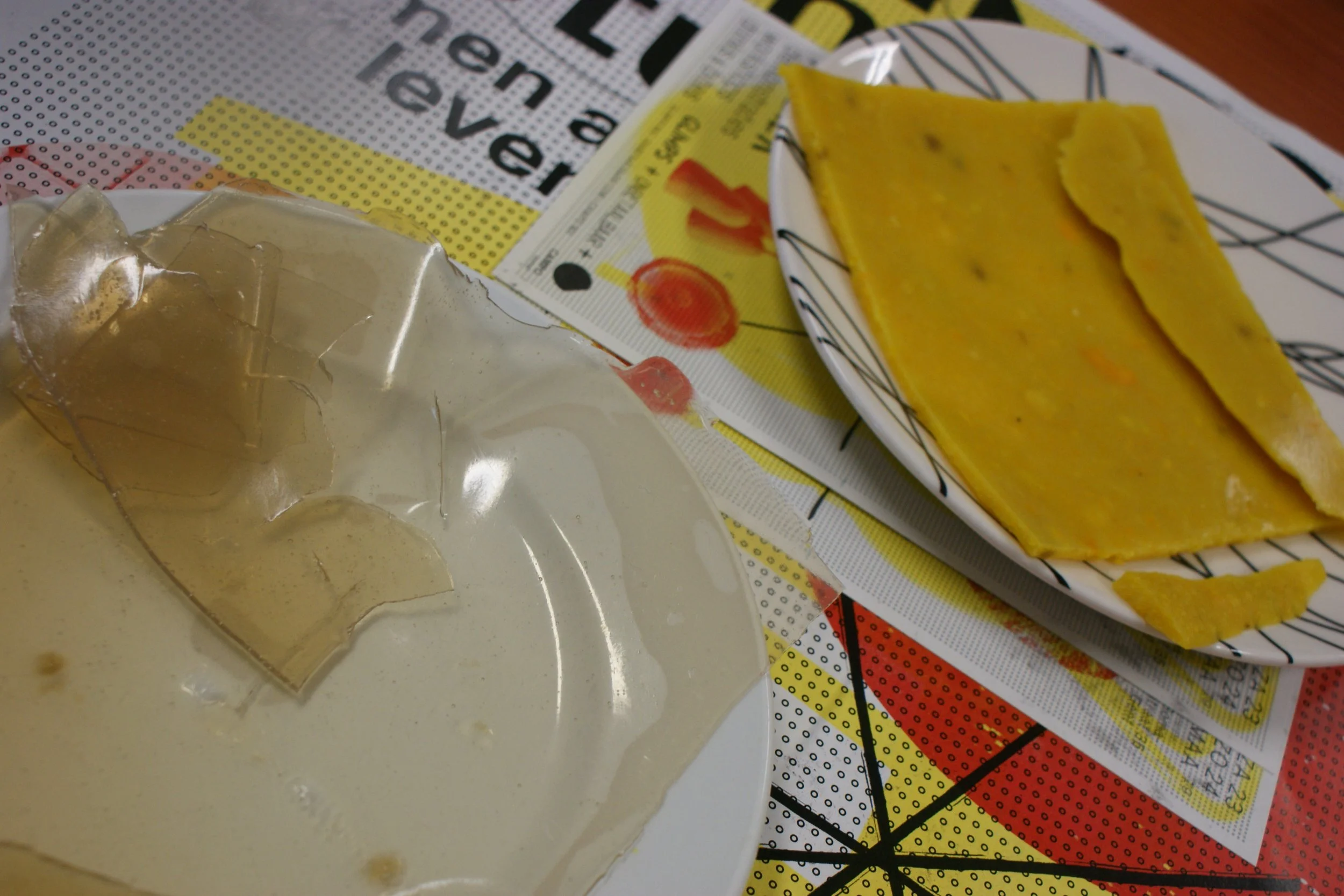 A partially unwrapped piece of butter or margarine, a plate with two pieces of yellow cheddar cheese, and some printed paper and magazines on a table.