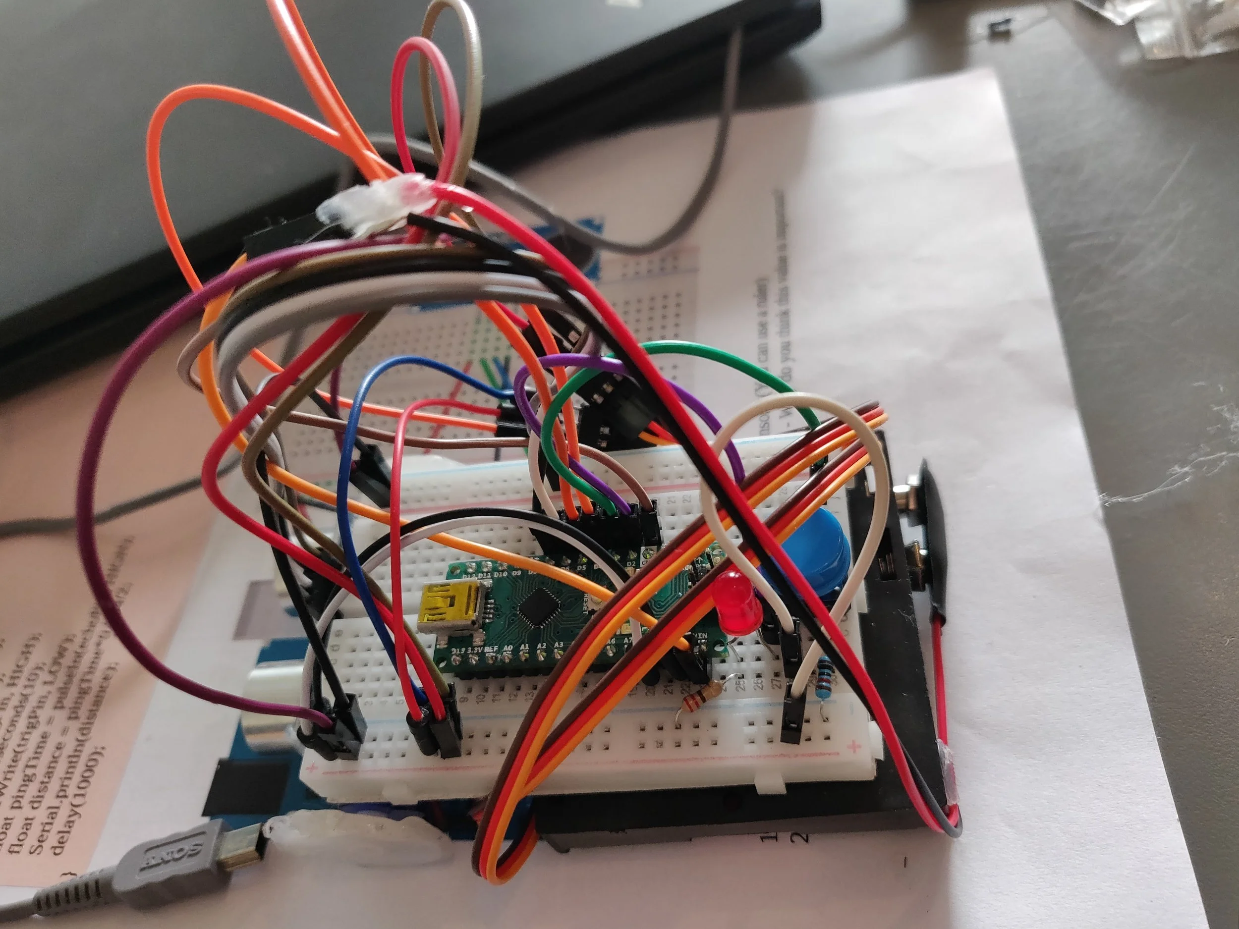 A breadboard with multiple colored wires connected to a small microcontroller, electronic components, and a USB power source, arranged on a paper surface.