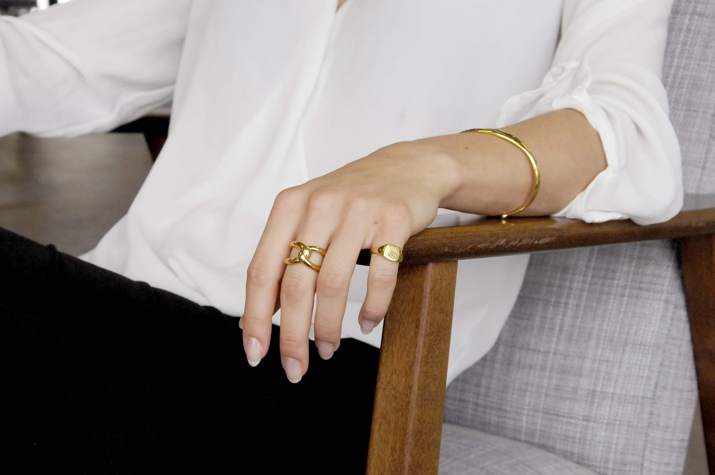 A jewellery model image of woman wearing gold jewellery, including rings, signet and bracelets, resting her hand on a wooden chair.