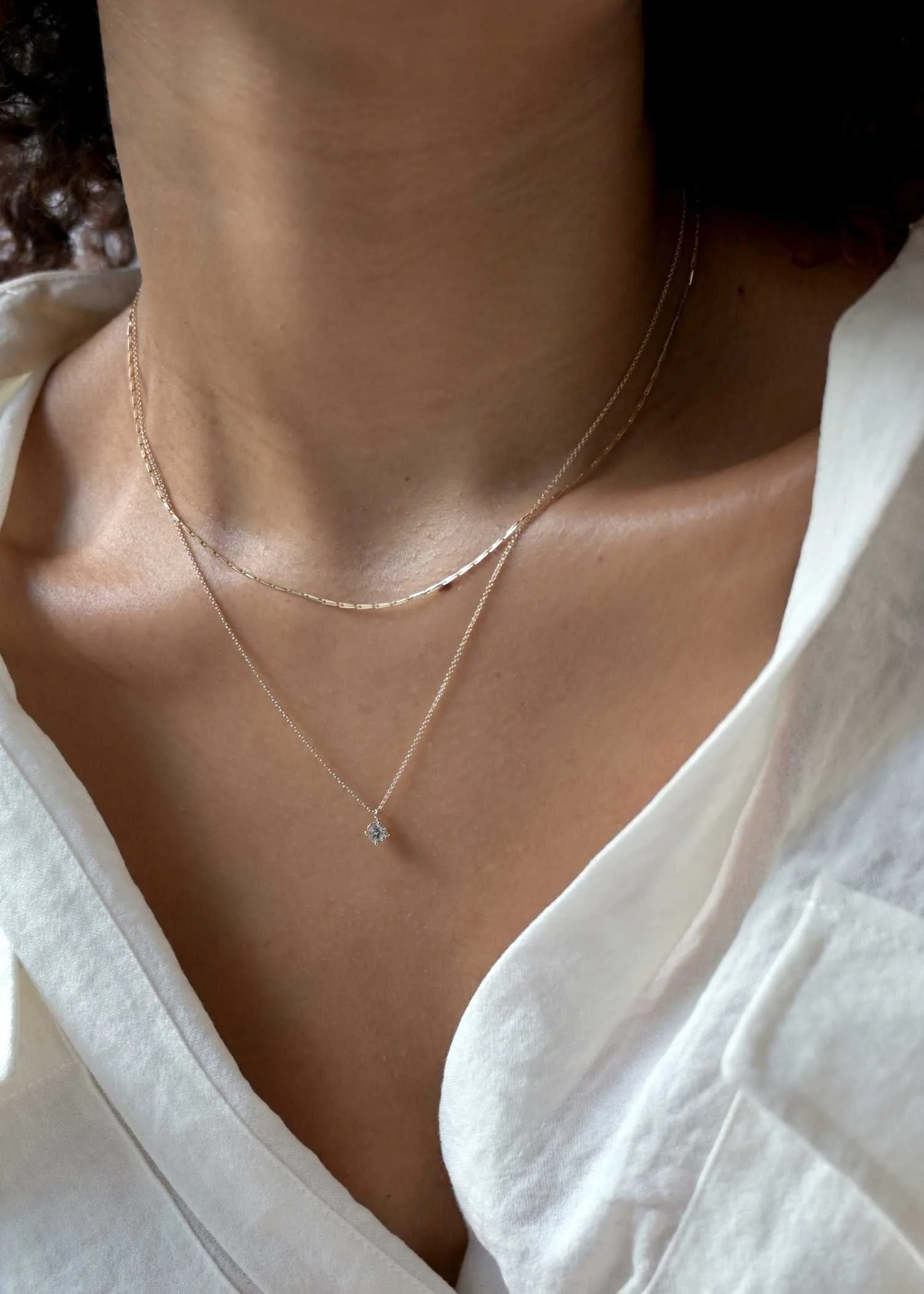Close-up of a woman's neck and collarbone area with layered gold necklaces, one with a small pendant, wearing a white shirt.