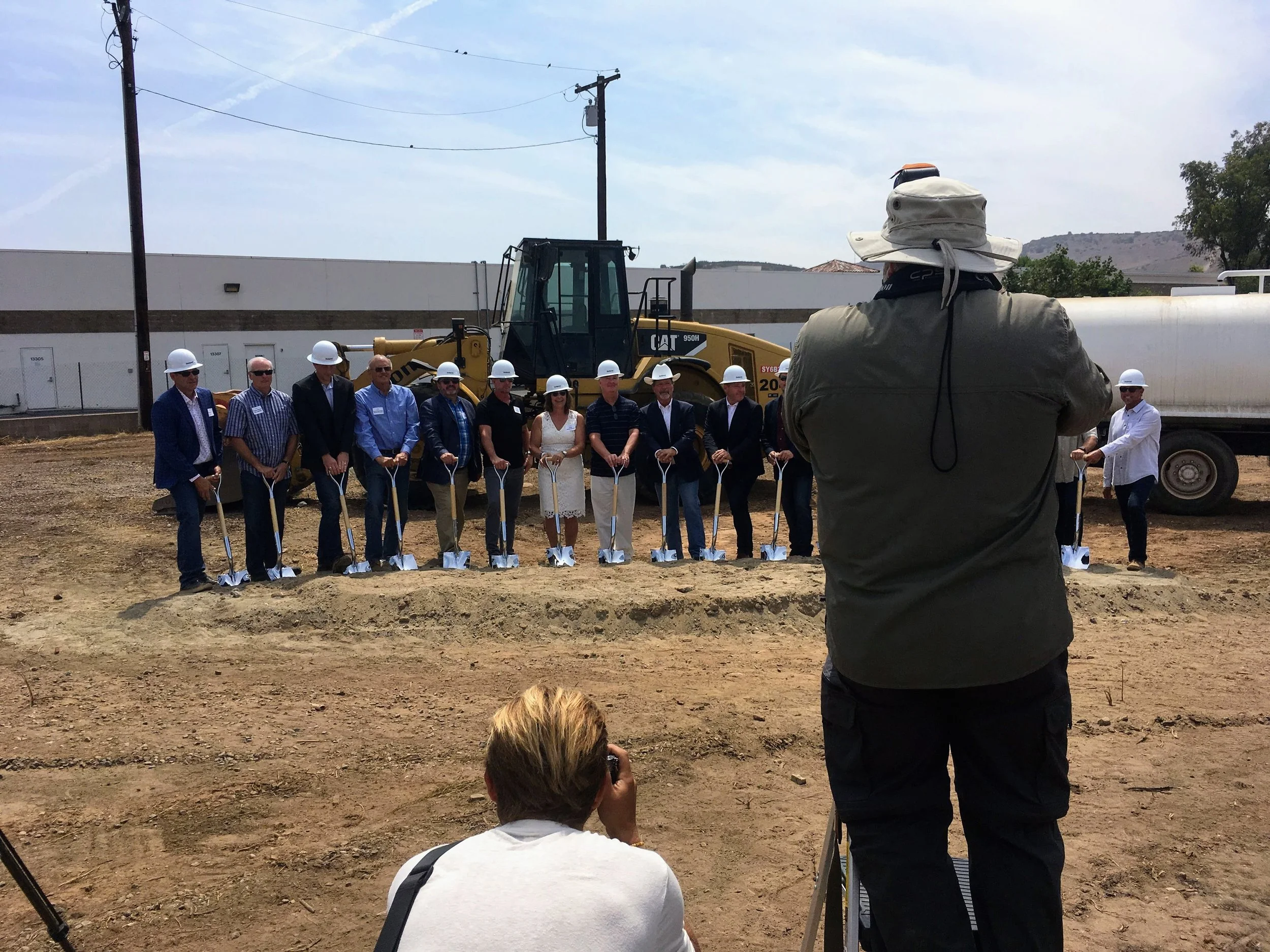 People wearing hard hats and holding shovels at groundbreaking while media films them for the news. 