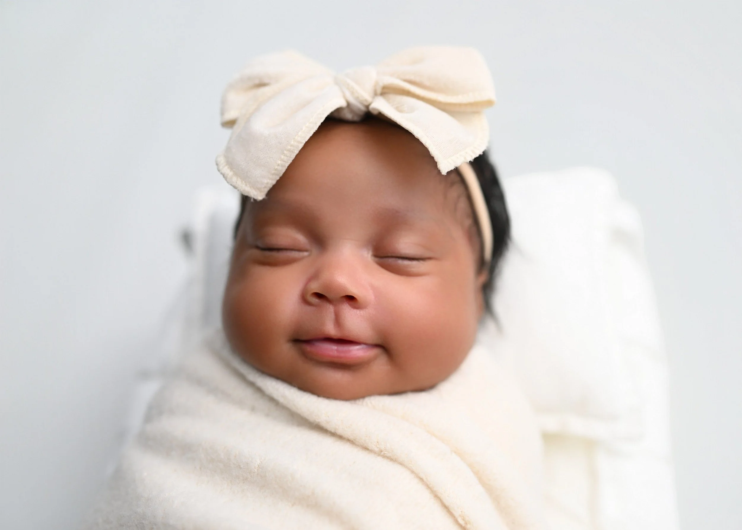 Close-up of a young child with closed eyes, smiling, wearing a white headband with a bow and a cream-colored shirt, against a plain light background.