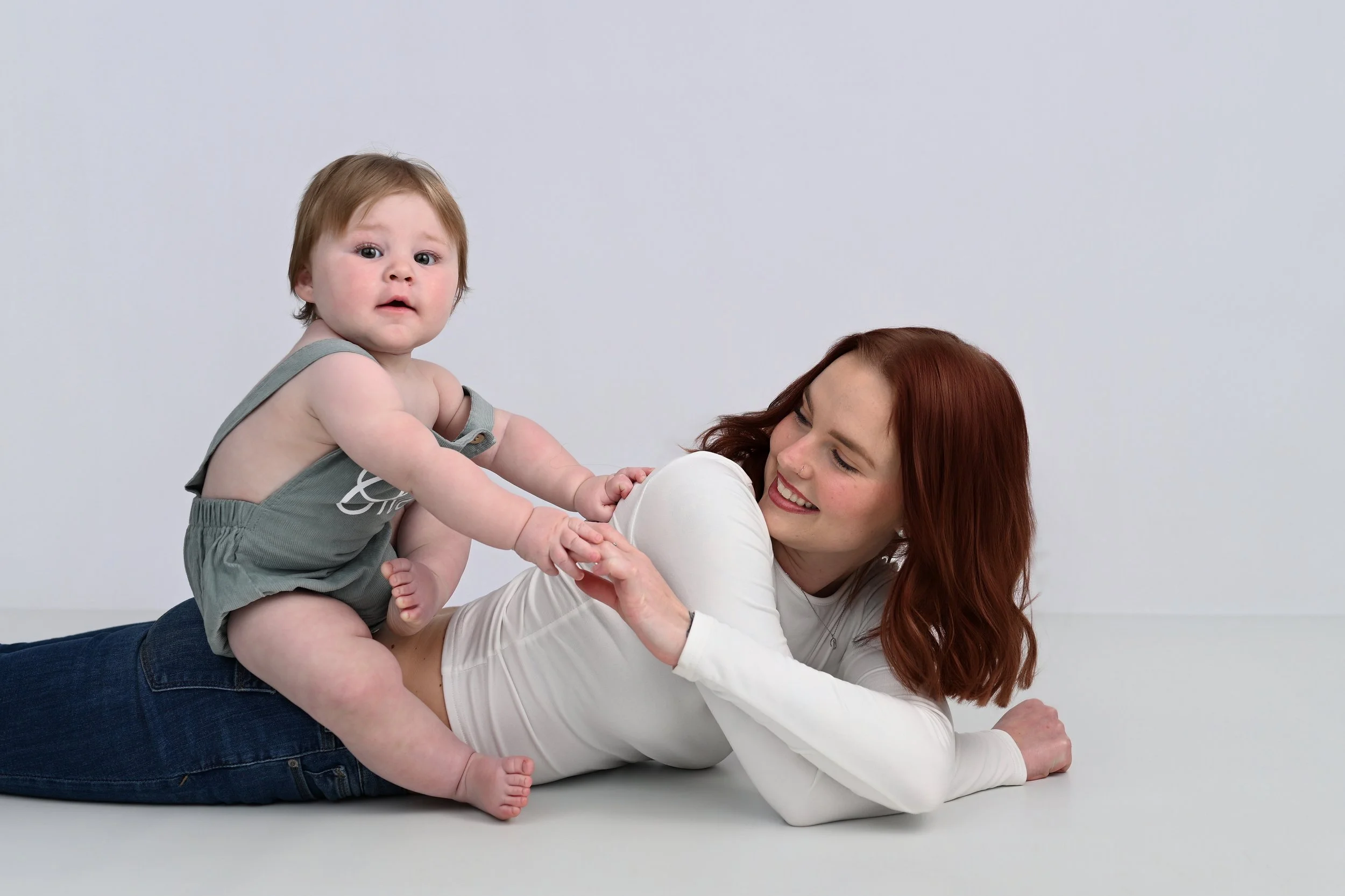 A woman with red hair and a white long-sleeve shirt lying on the floor with a big smile, holding a young child with light brown hair and light skin, wearing a grey sleeveless top and grey shorts, against a plain white background.