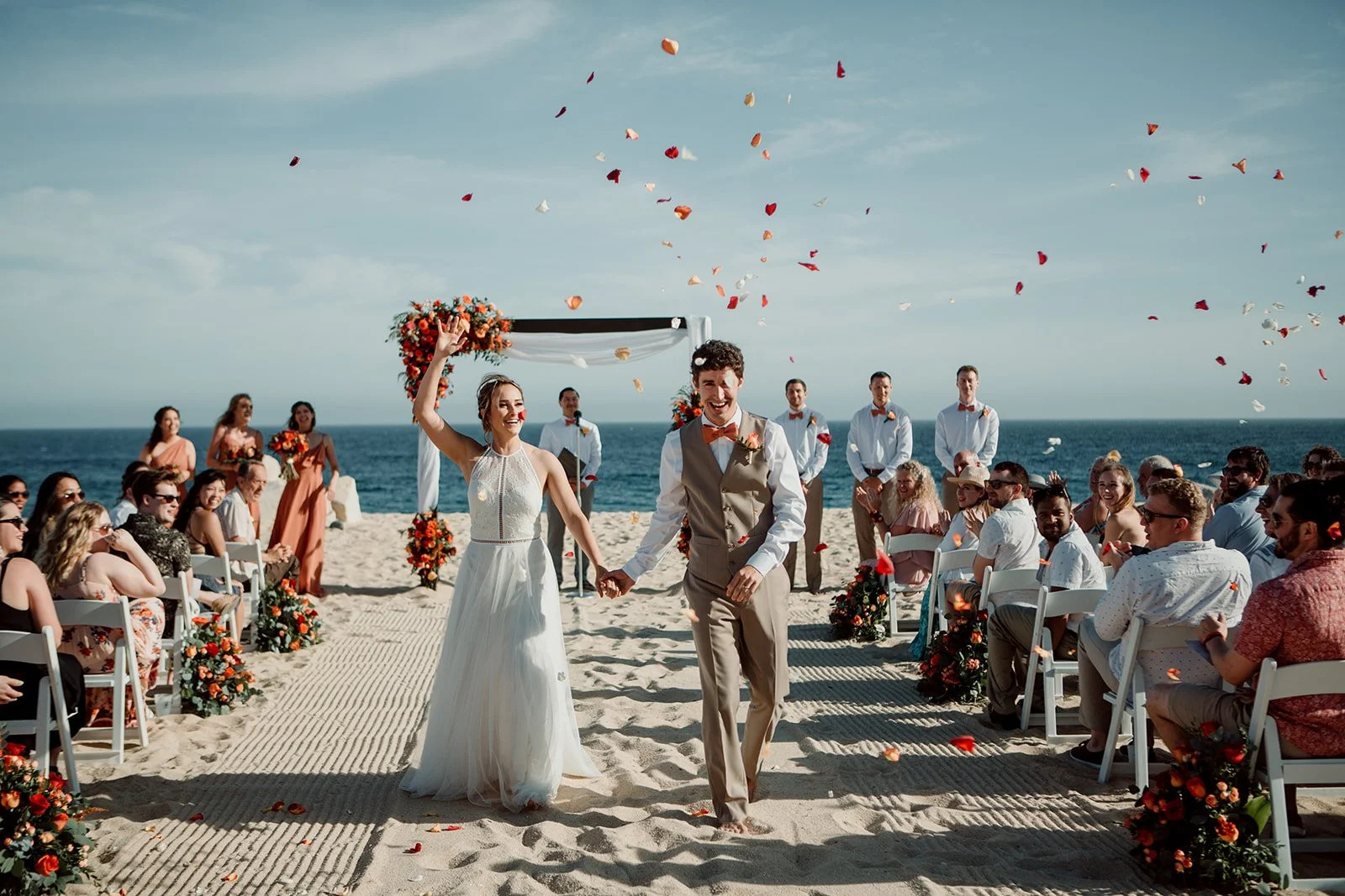 San Diego couple smiles holding hands while wedding guests throw colorful flower petals at them during their ceremony exit.