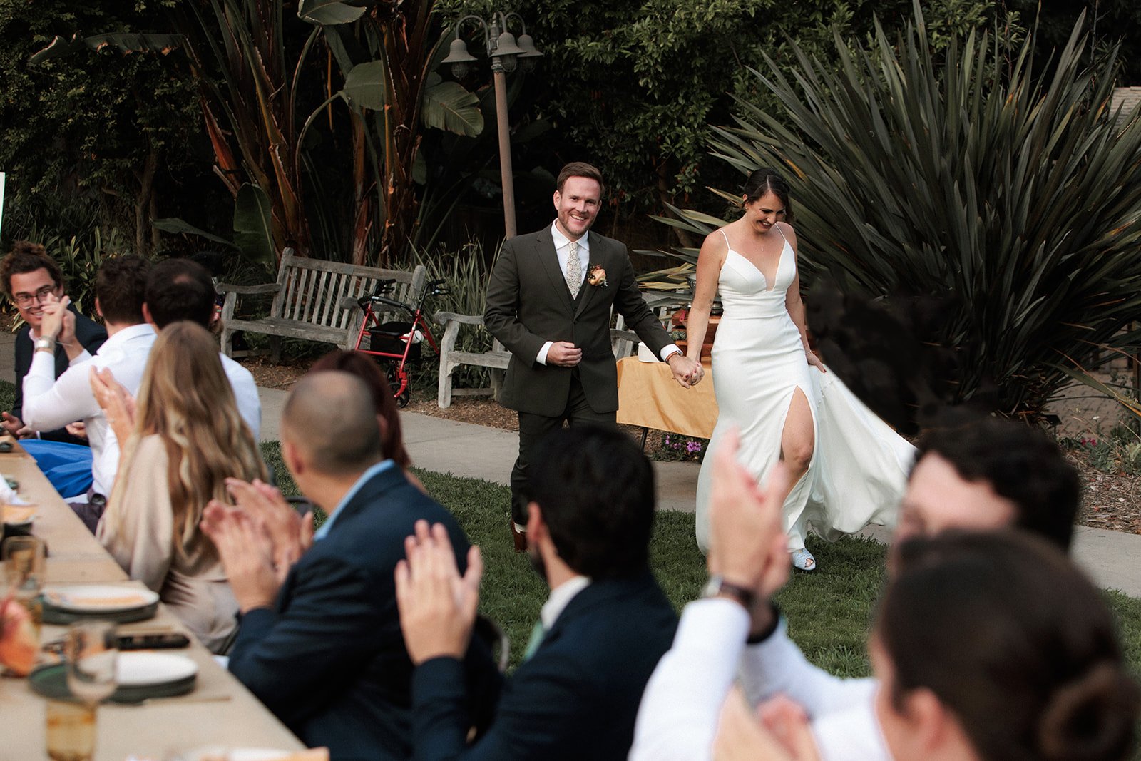 A couple smiles while waking into their grand entrance while guests look on and clap. San Diego Botanic Garden wedding.
