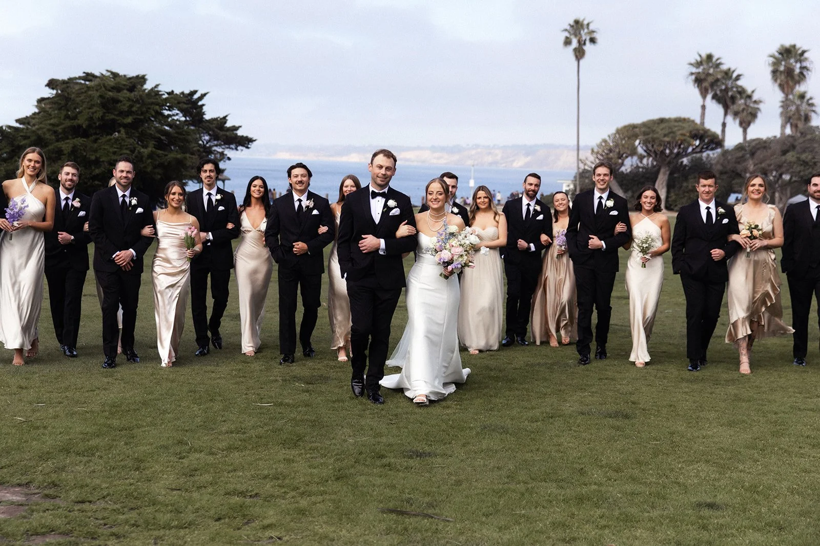 A couple walks across a grassy lawn overlooking La Jolla Cove with their wedding party behind them