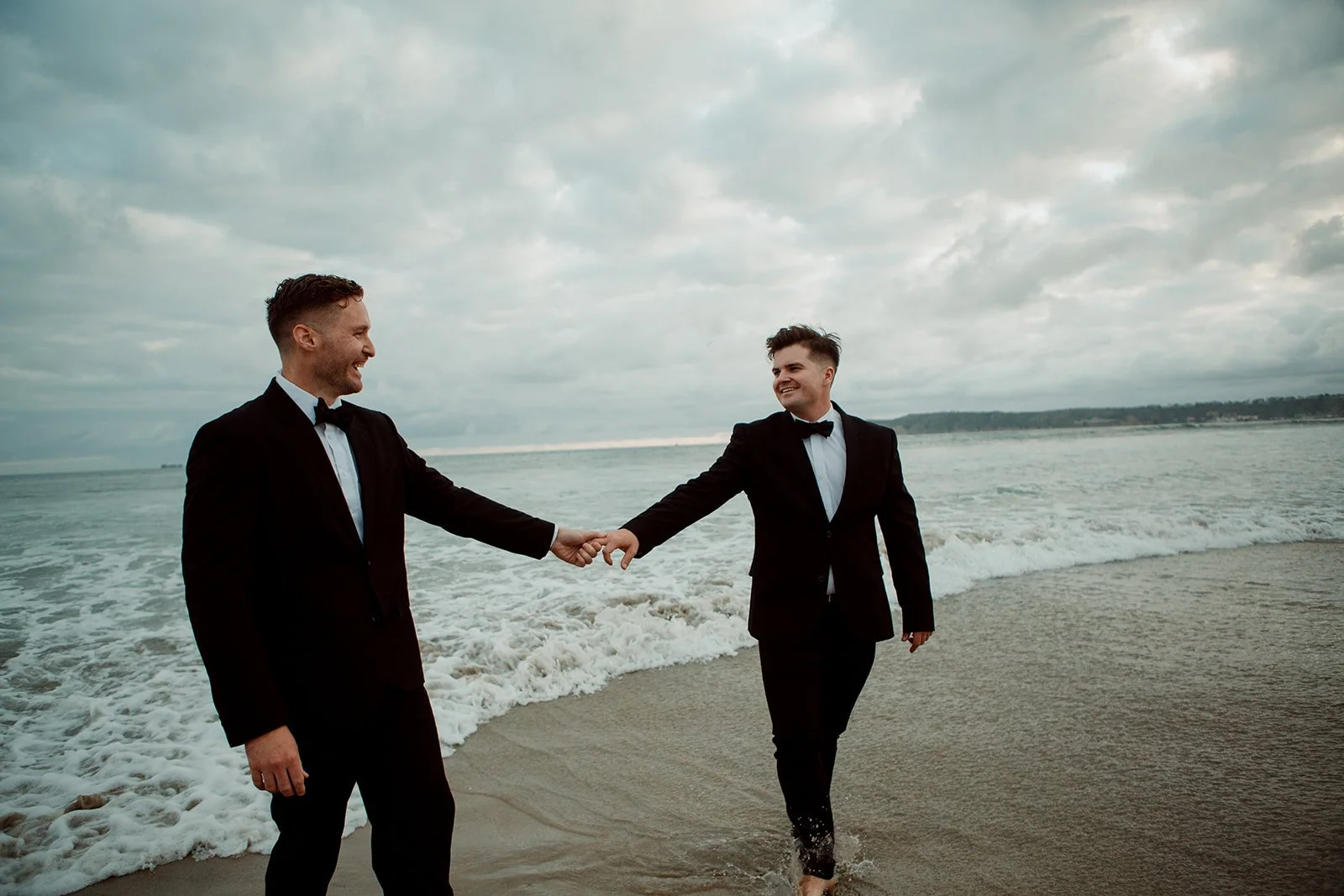 Two men hold hands and walk on the beach for their Coronado sunset engagement photo session.