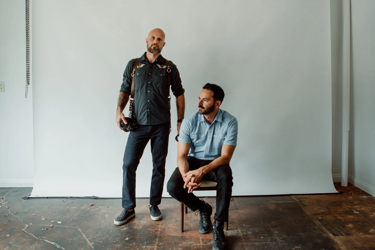 Photographers standing in their photography studio with a white backdrop. One man is standing with a camera, and the other is sitting on a chair, looking towards the standing man.
