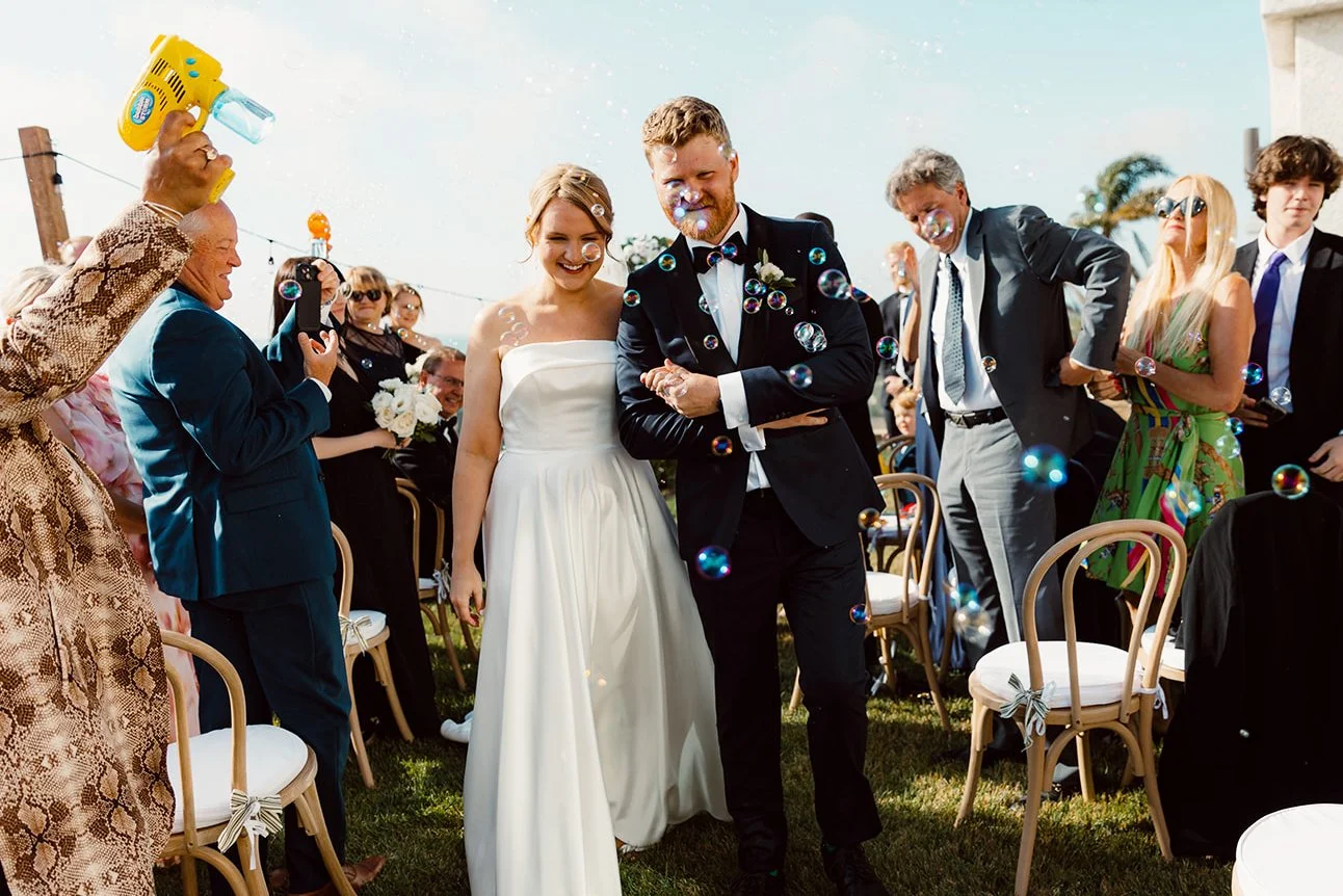 Bride and groom smile as wedding guests shoot bubbles towards them for their La Jolla wedding ceremony exit.