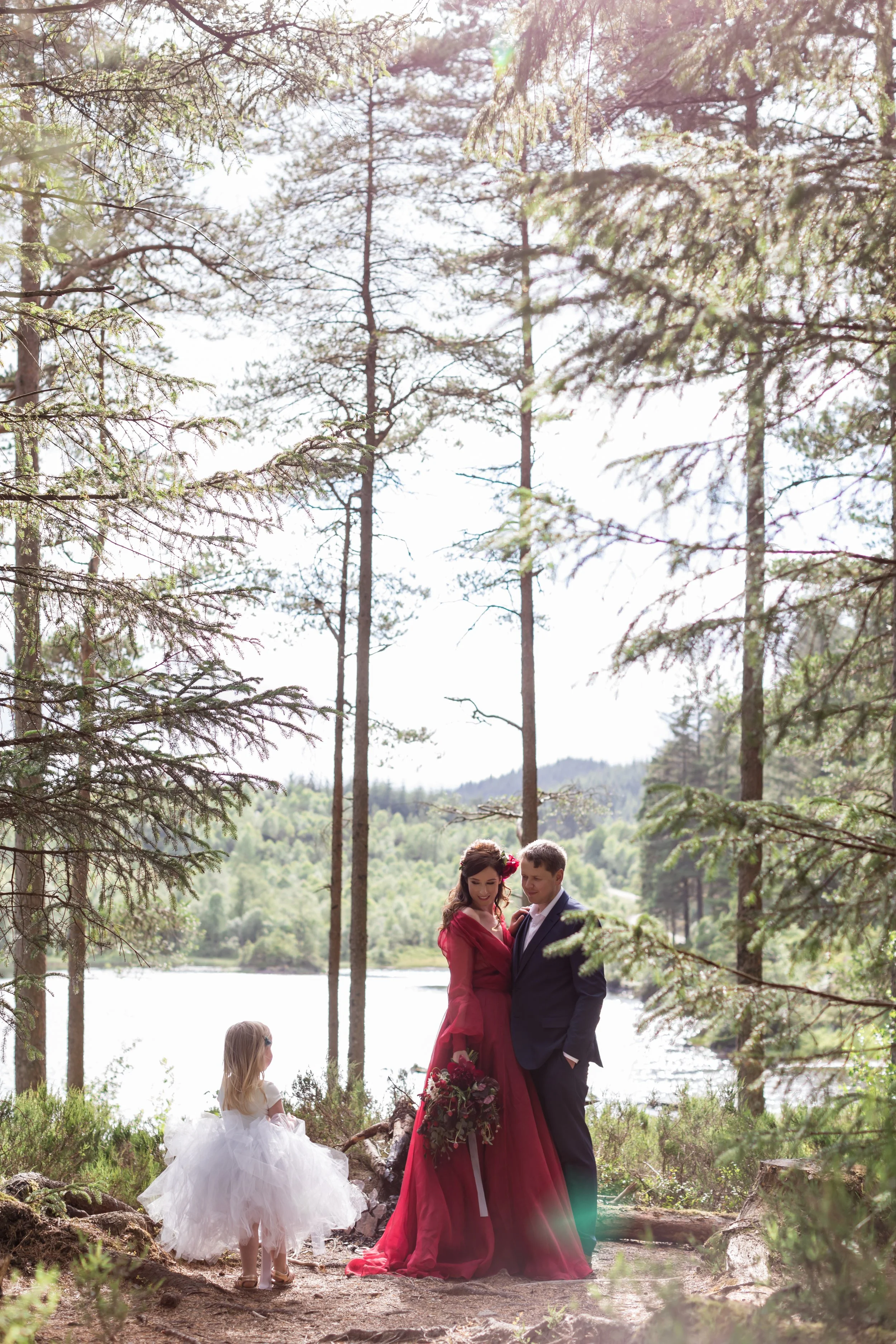 A couple dressed in wedding attire standing by a lake in a forest, with a young girl in a white dress nearby.