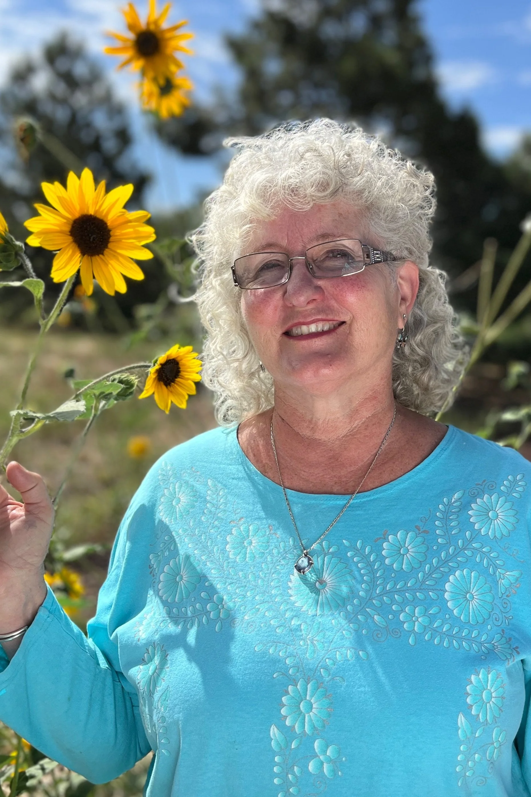 A smiling elderly woman with white curly hair, glasses, and earrings standing outdoors among yellow flowers, wearing a light blue top with floral embroidery, against a background of a blue sky and trees.