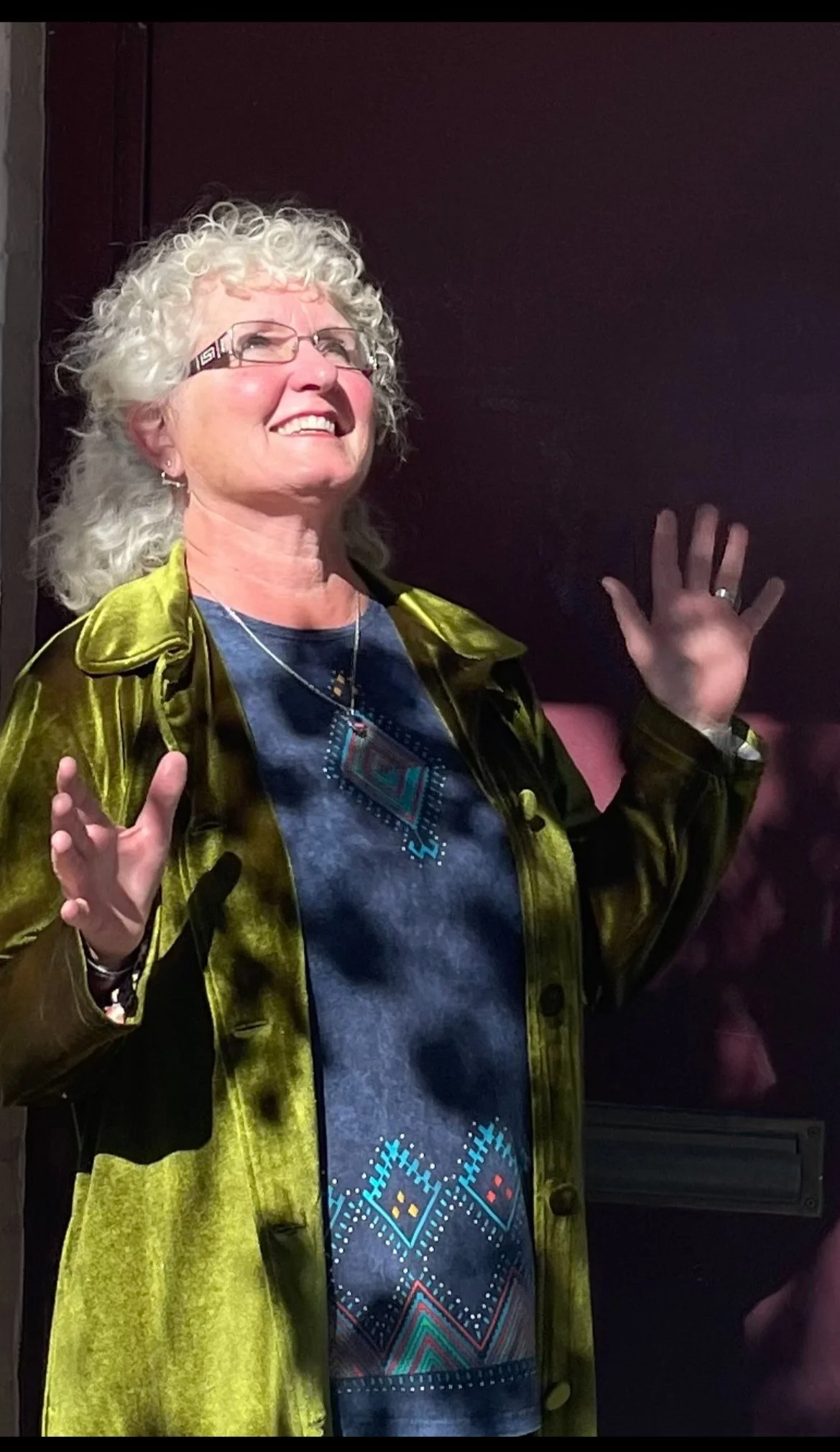 A woman with curly white hair, wearing glasses, a shiny green jacket, and a dark patterned shirt, smiling and raising her hands.
