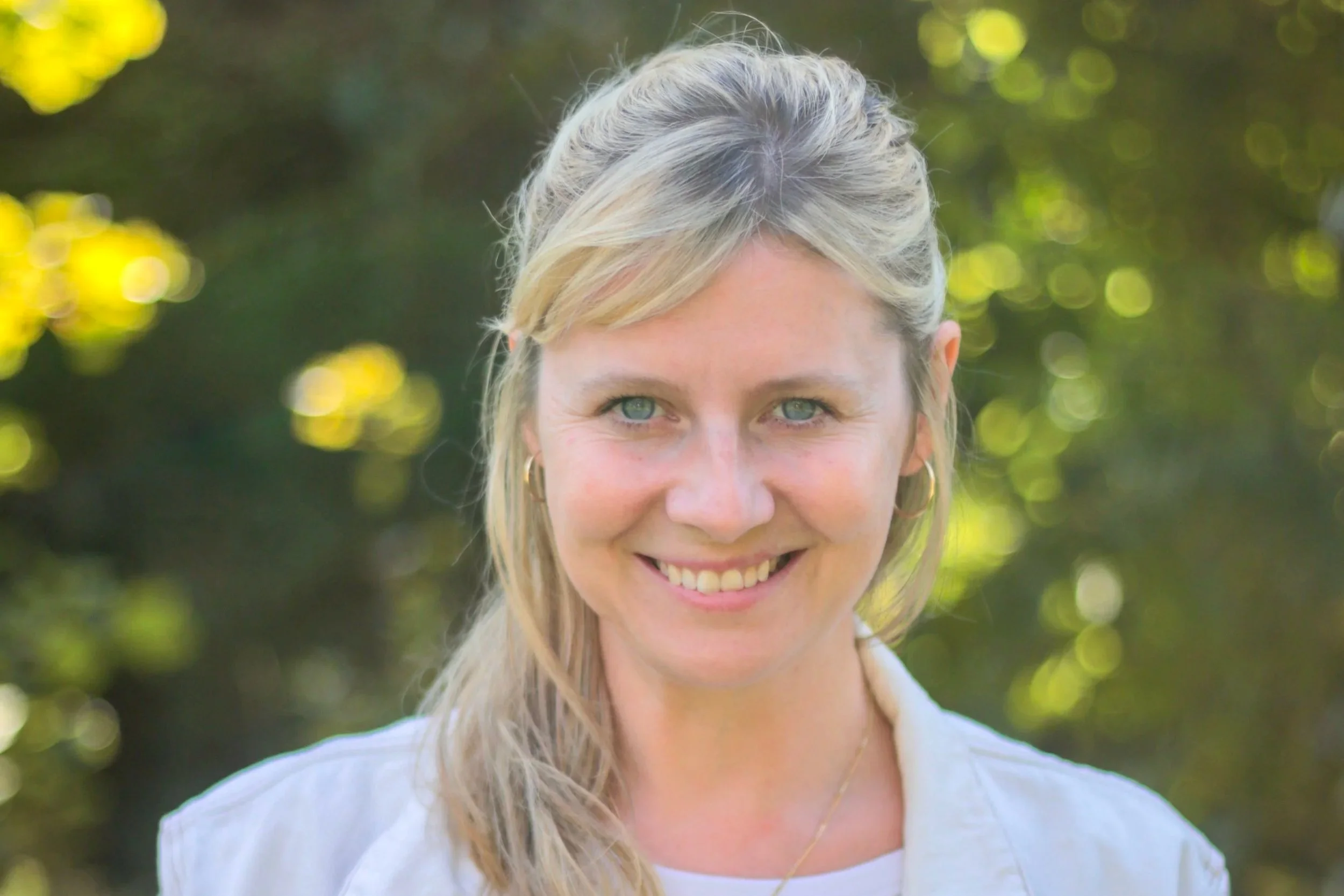Porträt einer lachenden Frau mit blondem Haar und blauen Augen vor unscharfem grünen Hintergrund.
