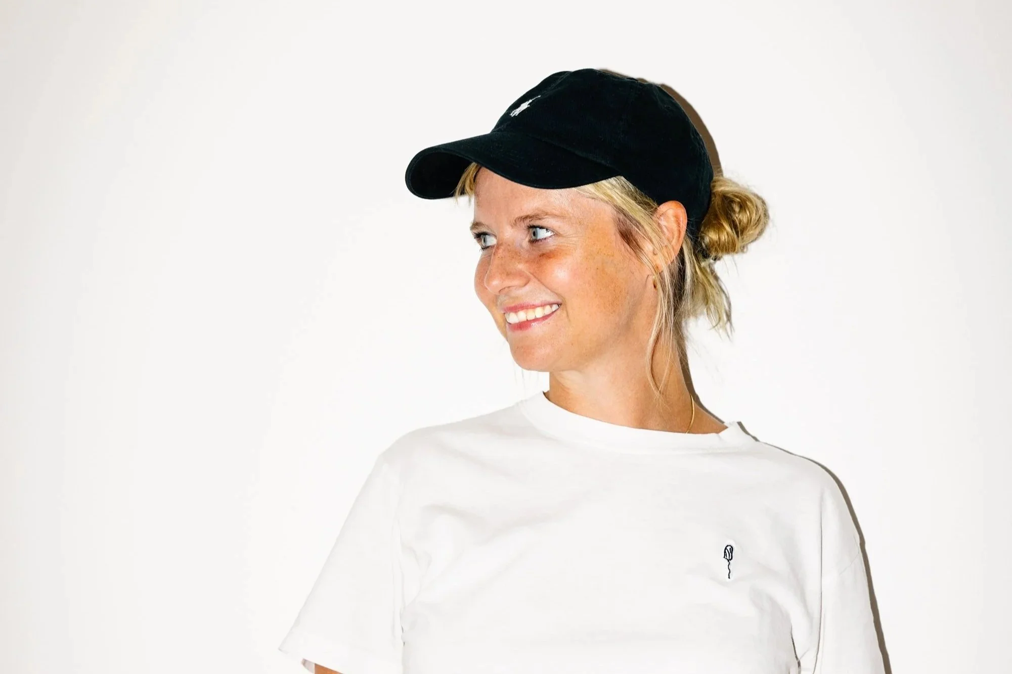 Junge Frau trägt schwarze Kappe und weißes T-Shirt, lächelt, blickt seitlich nach rechts, steht vor weißem Hintergrund.