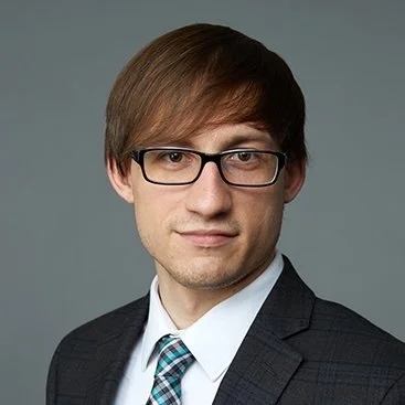 Professional headshot of a man with brown hair, glasses, wearing a suit and tie against a gray background.
