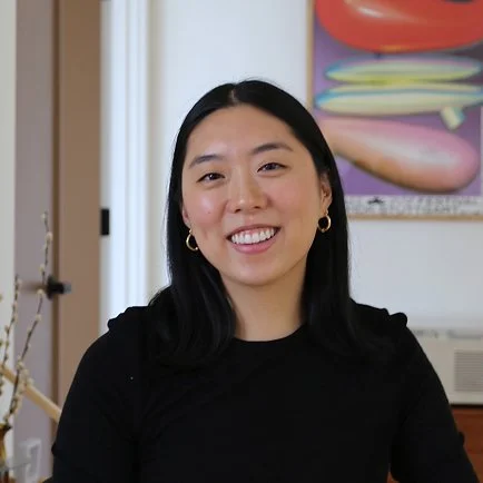 A young woman with black hair and gold hoop earrings smiling indoors with colorful artwork in the background.