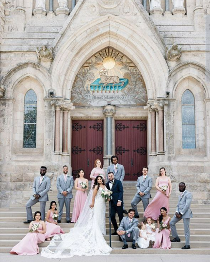 A wedding party poses in front of an ornate cathedral entrance. The bride, in a white gown, and the groom, in a dark suit, stand at the center. Bridesmaids in pink dresses and groomsmen in gray suits surround them, some sitting and some standing on the steps. The architecture features large arched doors and stained glass windows.