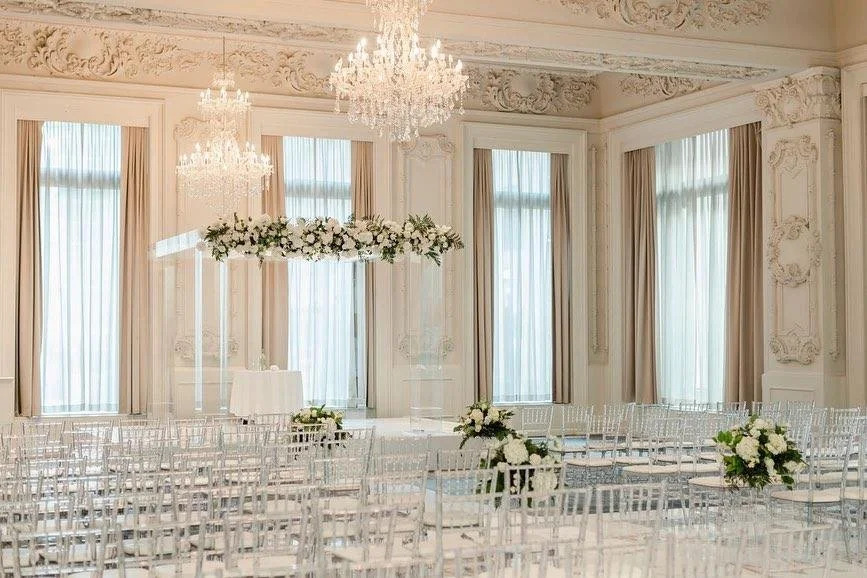 Elegant wedding ceremony setup in a luxurious room with chandeliers, floral arrangements, clear chairs, and ornate decorations.