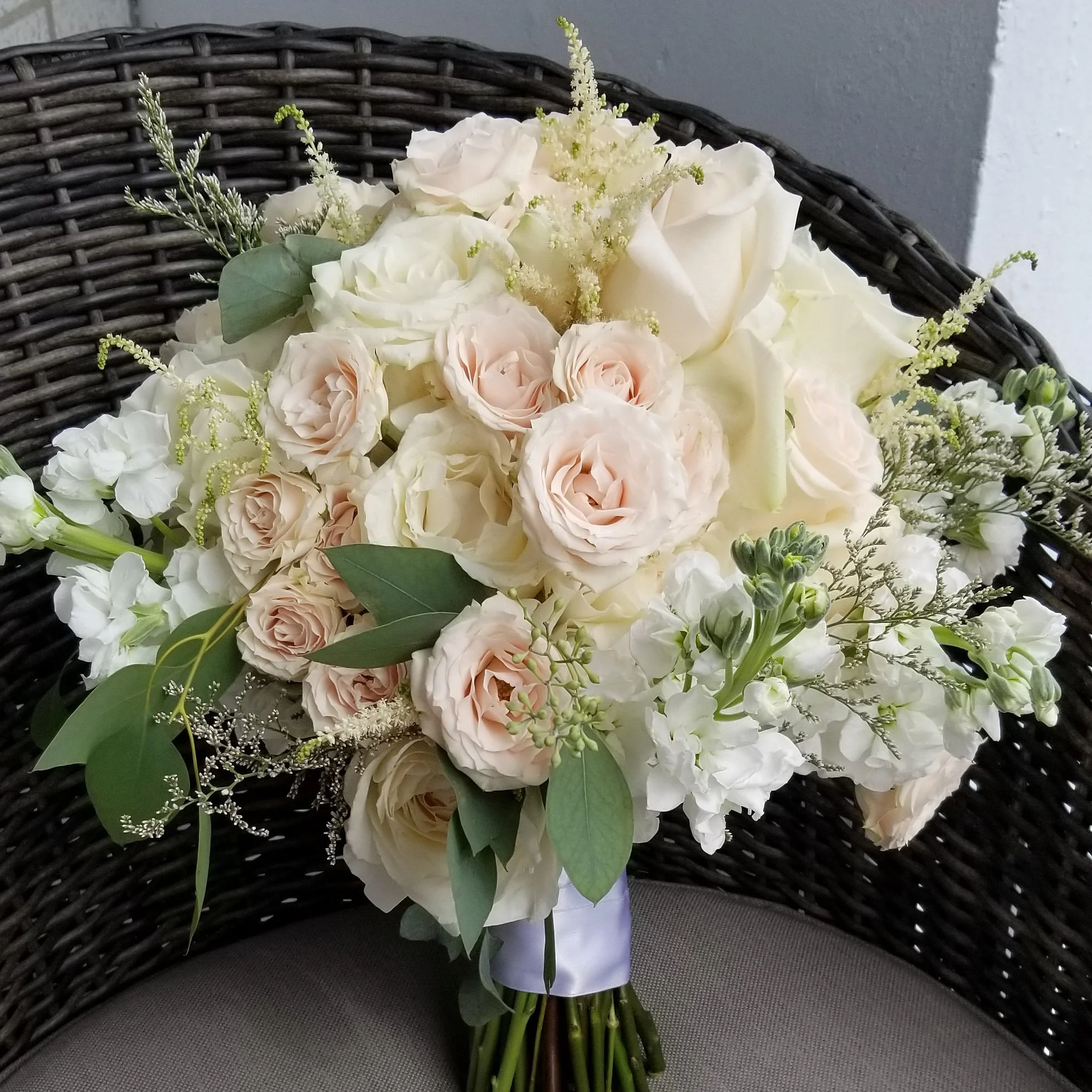 Bridal bouquet with white and pale pink roses, eucalyptus leaves, and other white flowers on a wicker chair.